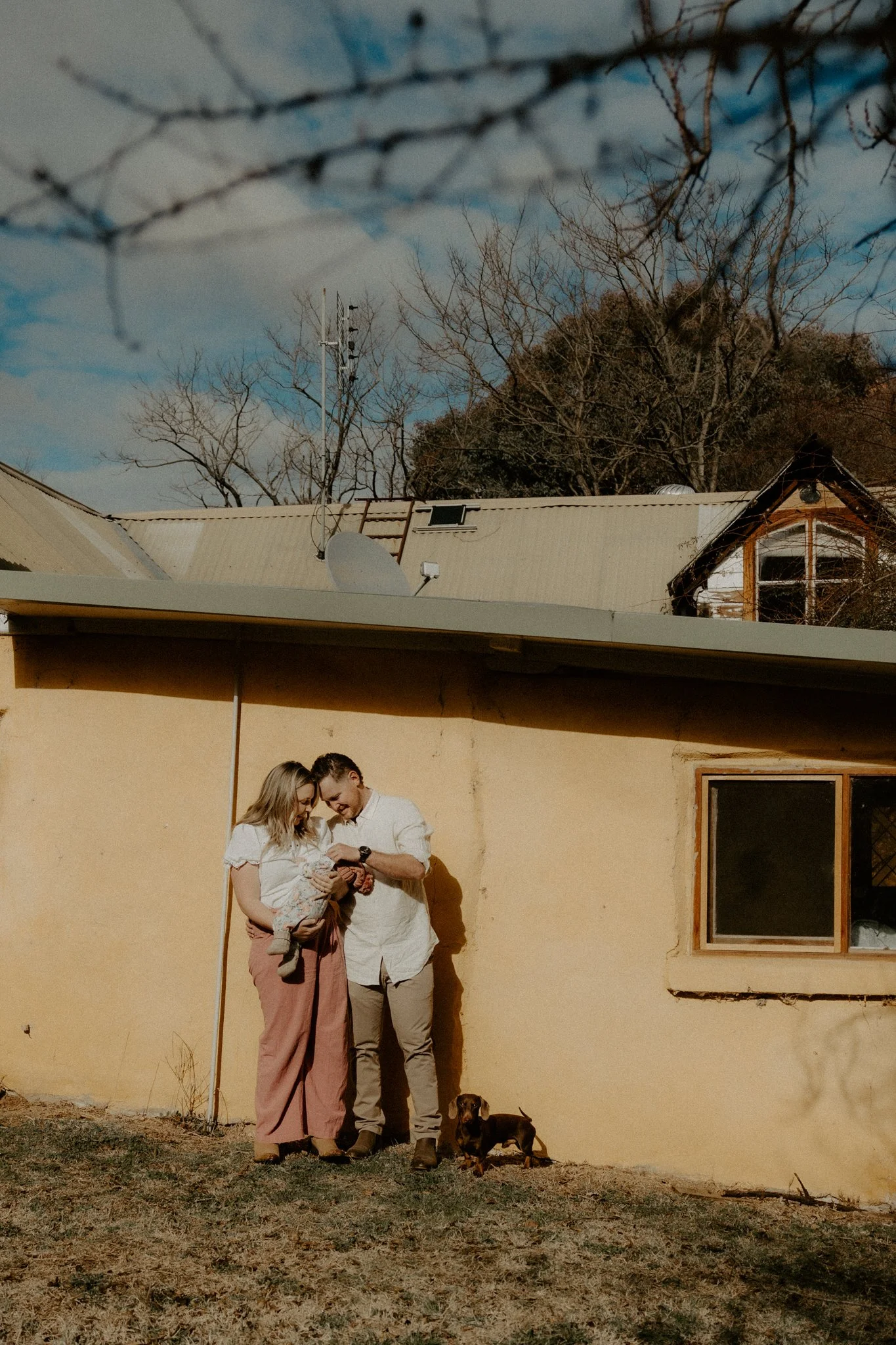 A couple with a child and a small dog standing outside near a beige wall of a house on a sunny day, looking at a device they are holding.