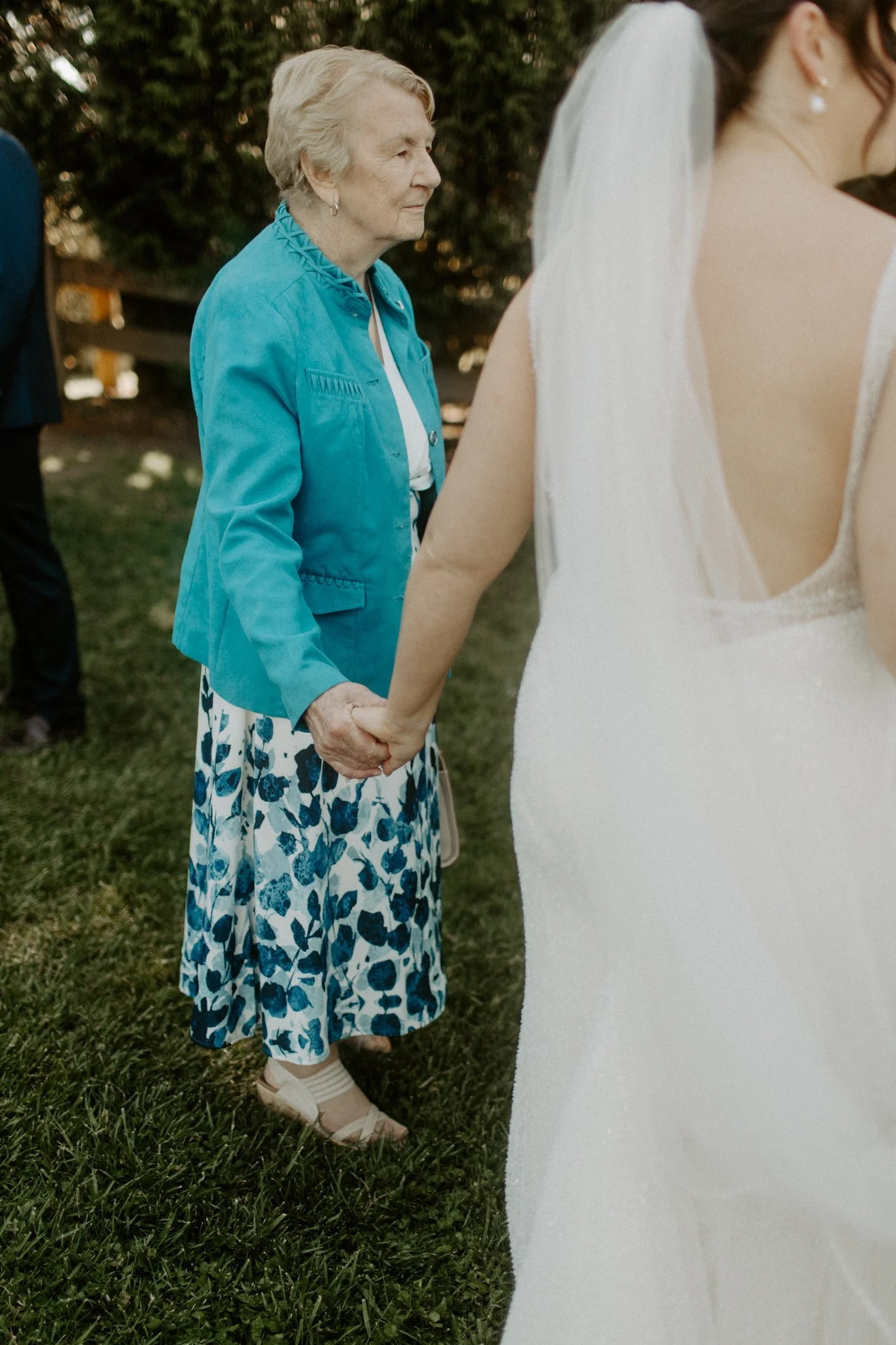 An elderly woman and a bride holding hands outdoors at a wedding ceremony.
