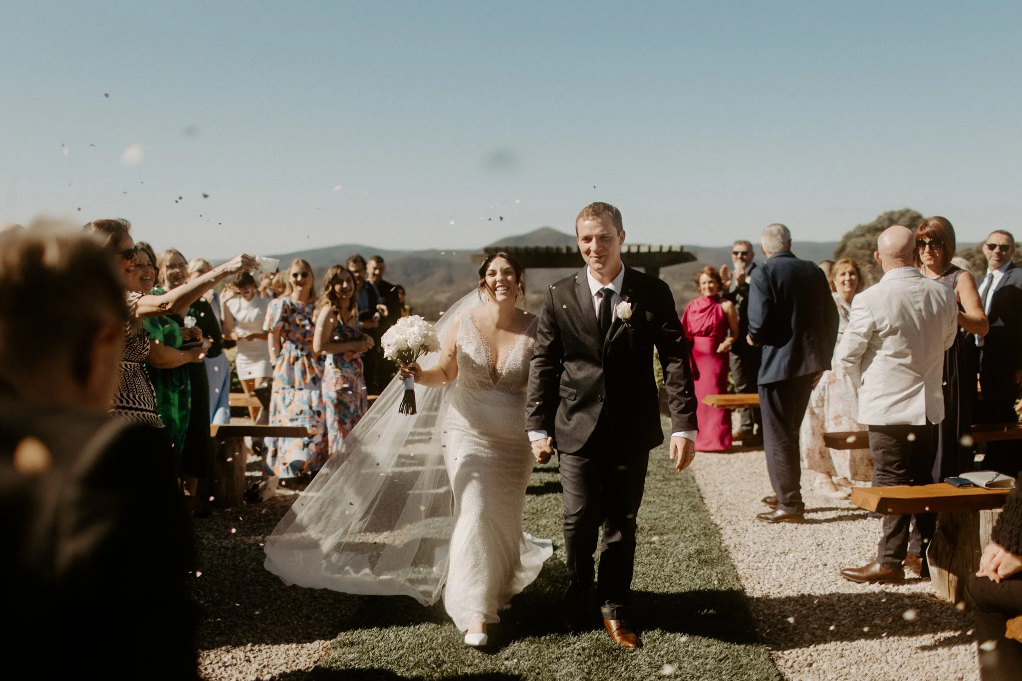 A bride and groom walking hand in hand down an outdoor aisle during a wedding ceremony, surrounded by cheering guests during a Blue Mountains wedding at Seclusions Blue Mountains.