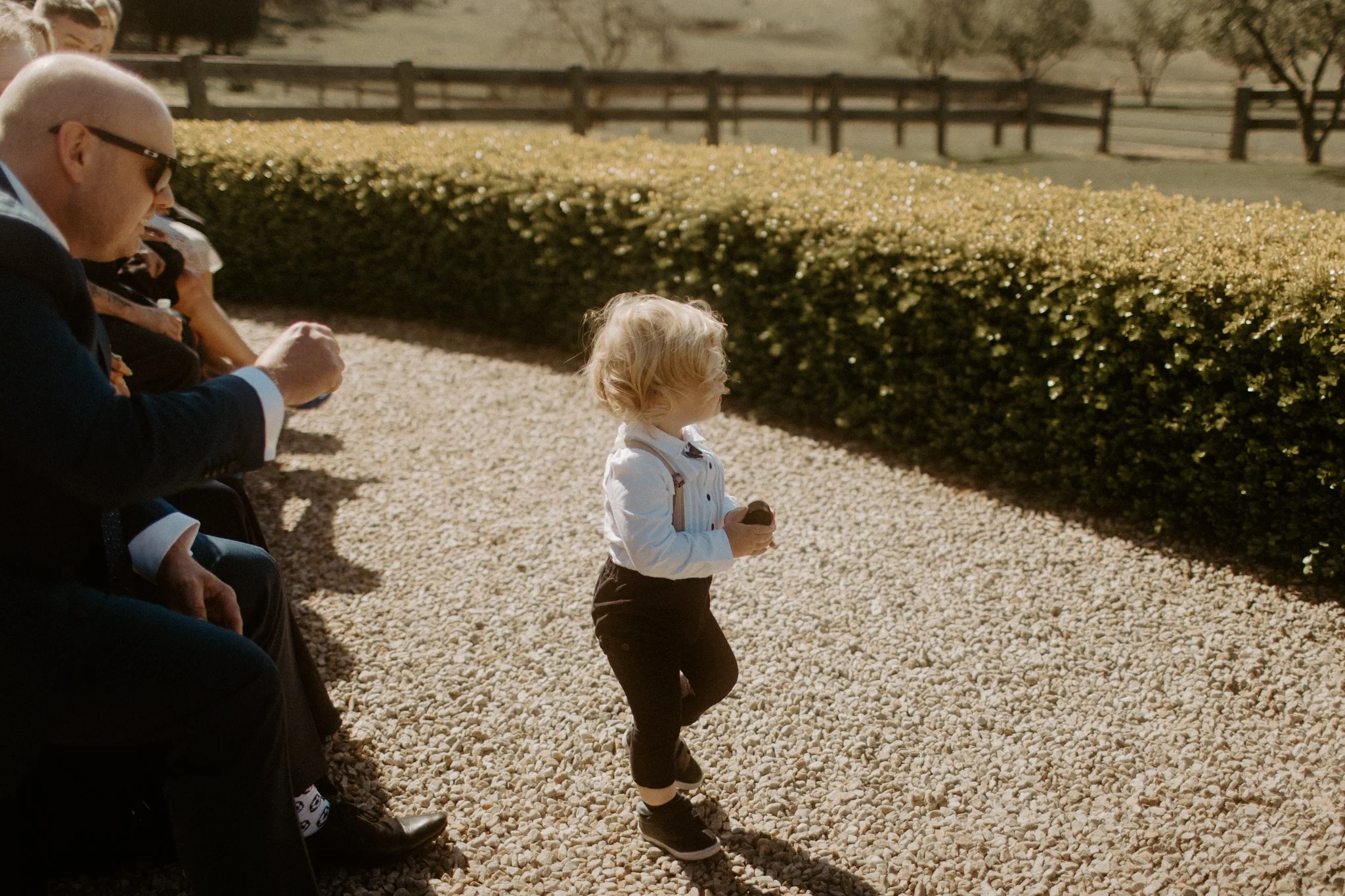 A young boy with curly blonde hair, wearing a white shirt and black pants, standing on a gravel path and holding a cookie. He is looking to the right. To the left, several adults are sitting and wearing sunglasses, also holding cookies. There is a ne