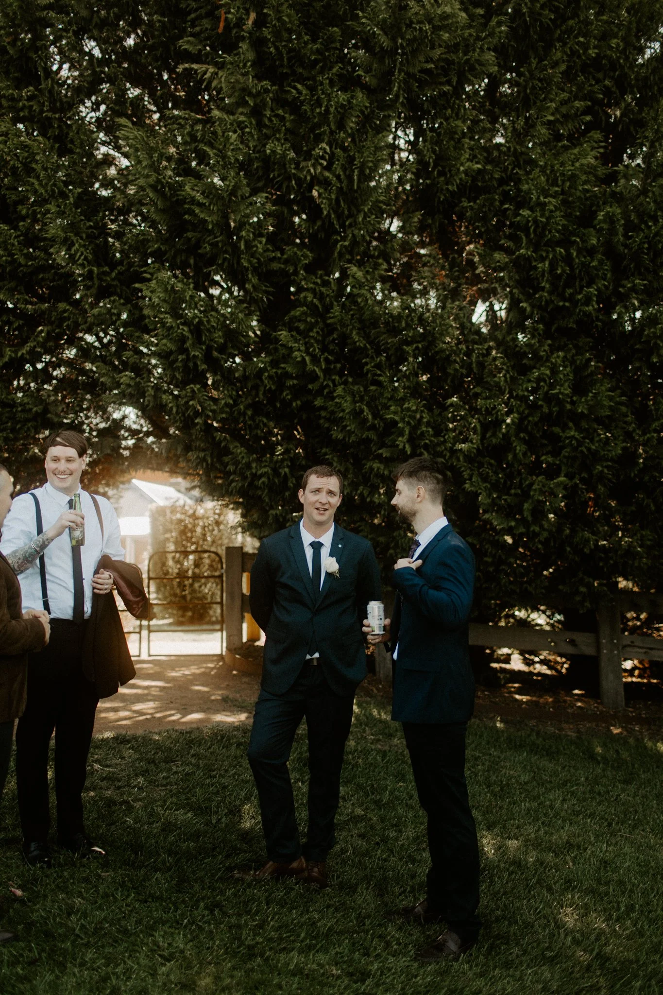 Group of men in suits talking outdoors near large evergreen trees, with one person smiling and another holding a beer bottle.