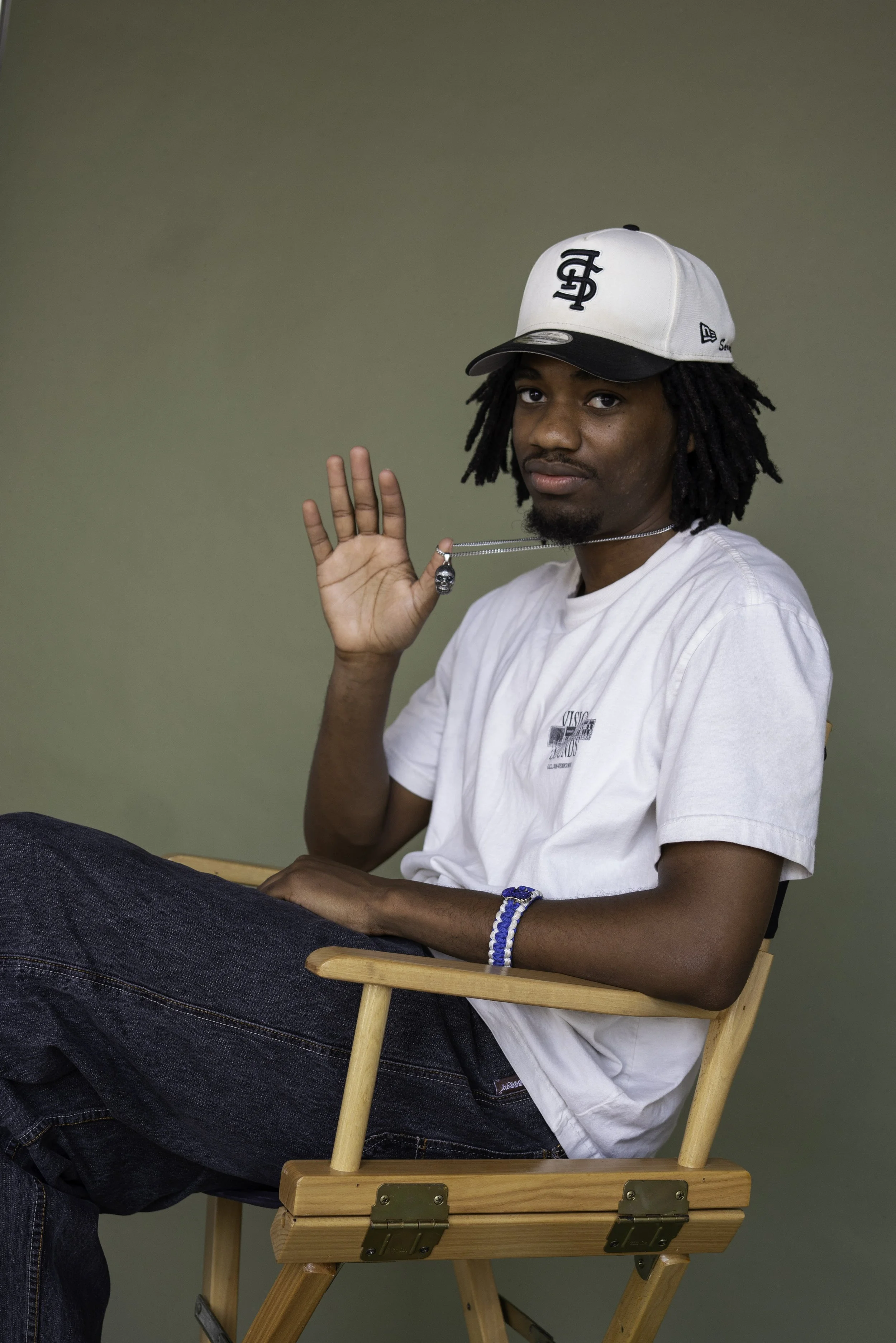 A young man with dreadlocks sitting on a wooden chair, wearing a white baseball cap with a black logo, a white t-shirt, and dark jeans. He is holding a small skull necklace in his right hand, slightly raised, and looking at the camera.
