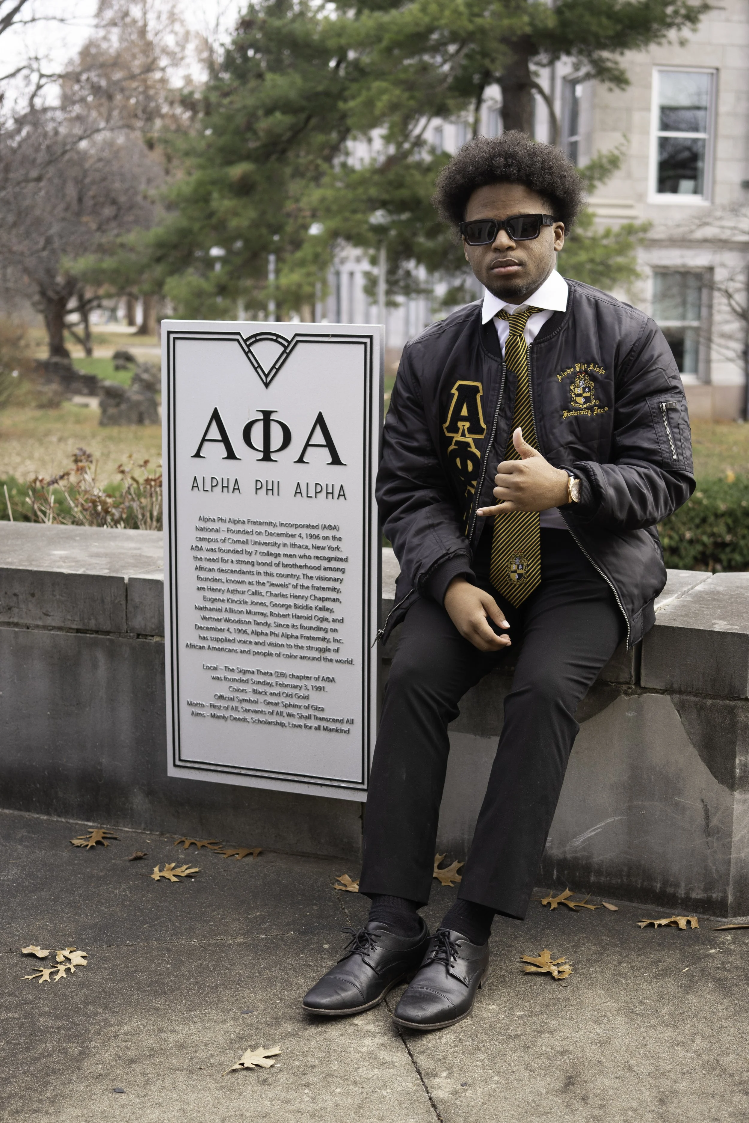 A young man dressed in black jeans, a black jacket with Alpha Phi Alpha letters, and a black and gold tie, sitting on a stone ledge beside an Alpha Phi Alpha fraternity informational sign outdoors in a park with leafless trees.
