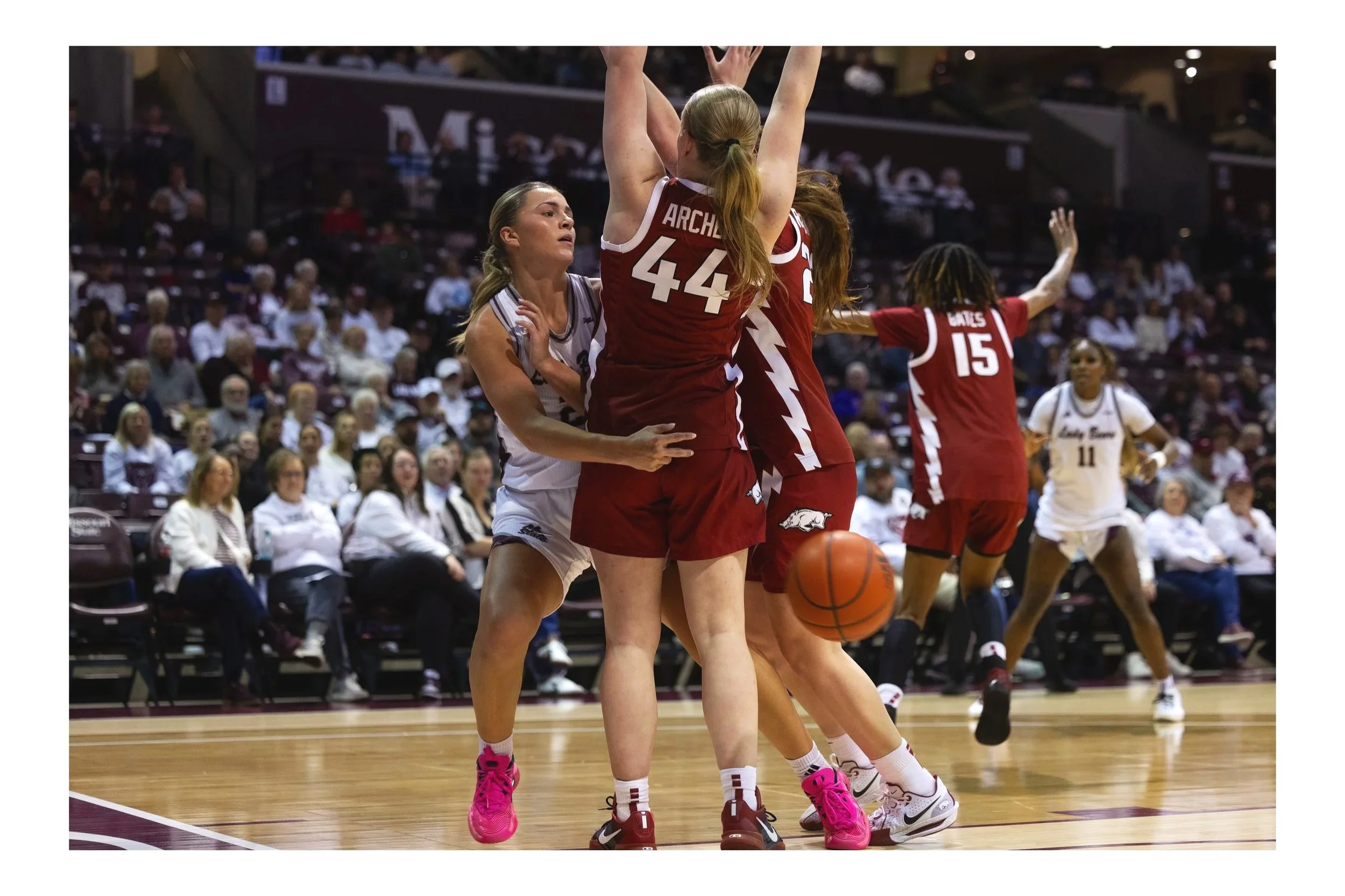 Women playing basketball in a game, with one woman in white trying to block a woman in red jumping to shoot or pass, with ARCHE visible on her jersey.