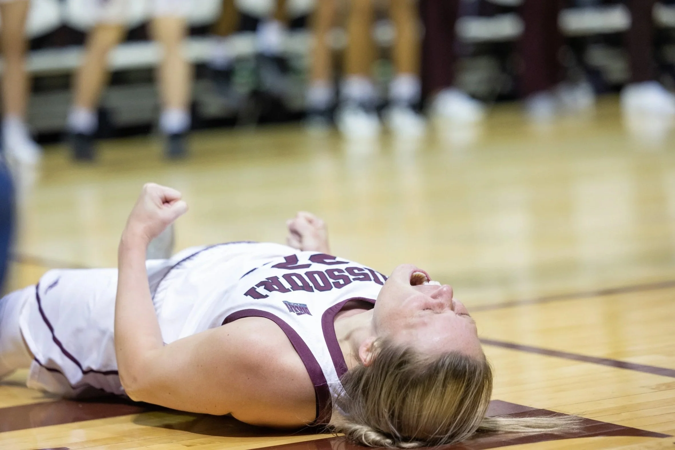 A female basketball player lies on the gym floor after what appears to be a fall or injury, with her face expressing pain.