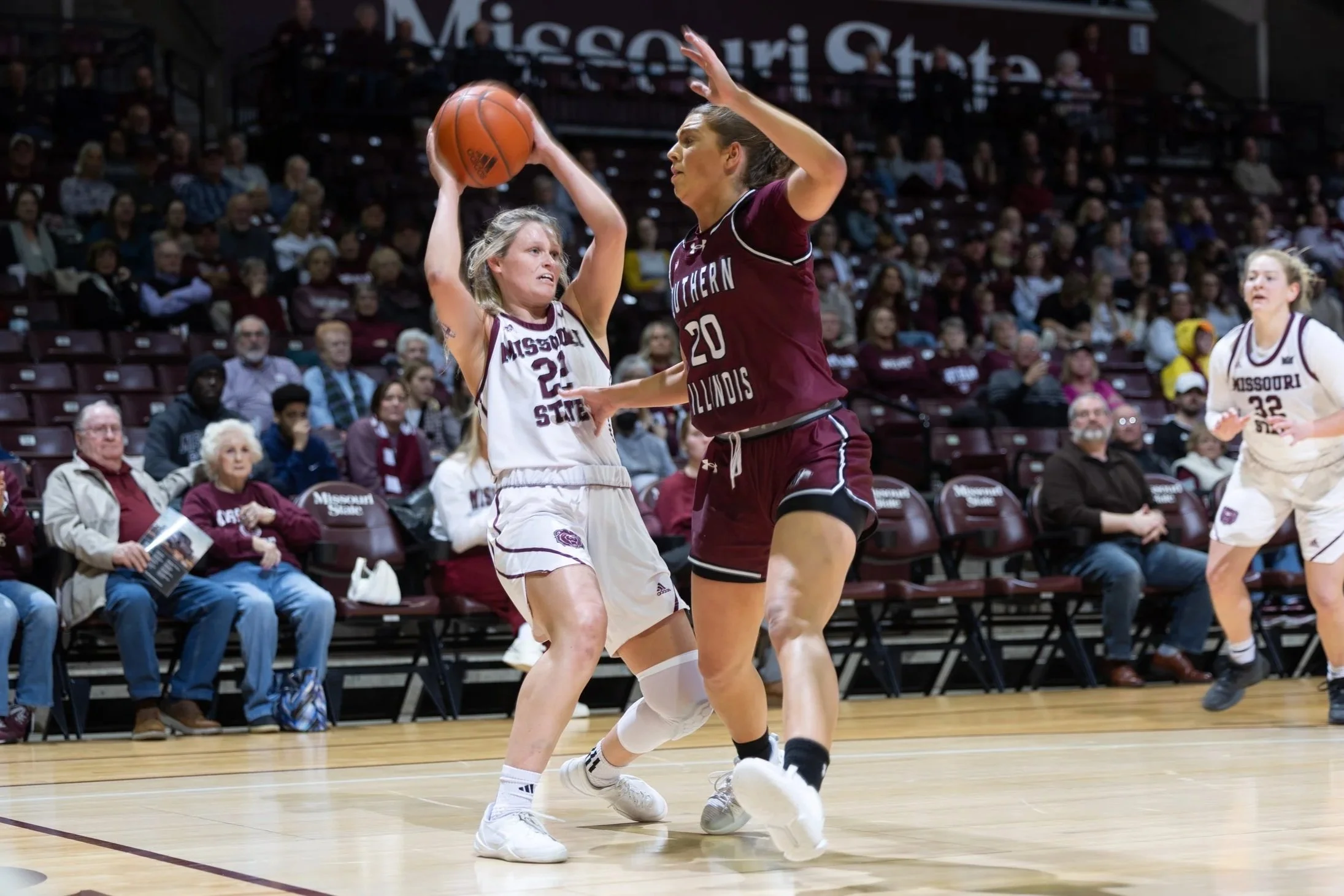 Two female basketball players, one in a white Missouri State uniform and the other in a maroon Southern Illinois uniform, compete for the basketball during a game. Spectators watch from the stands.