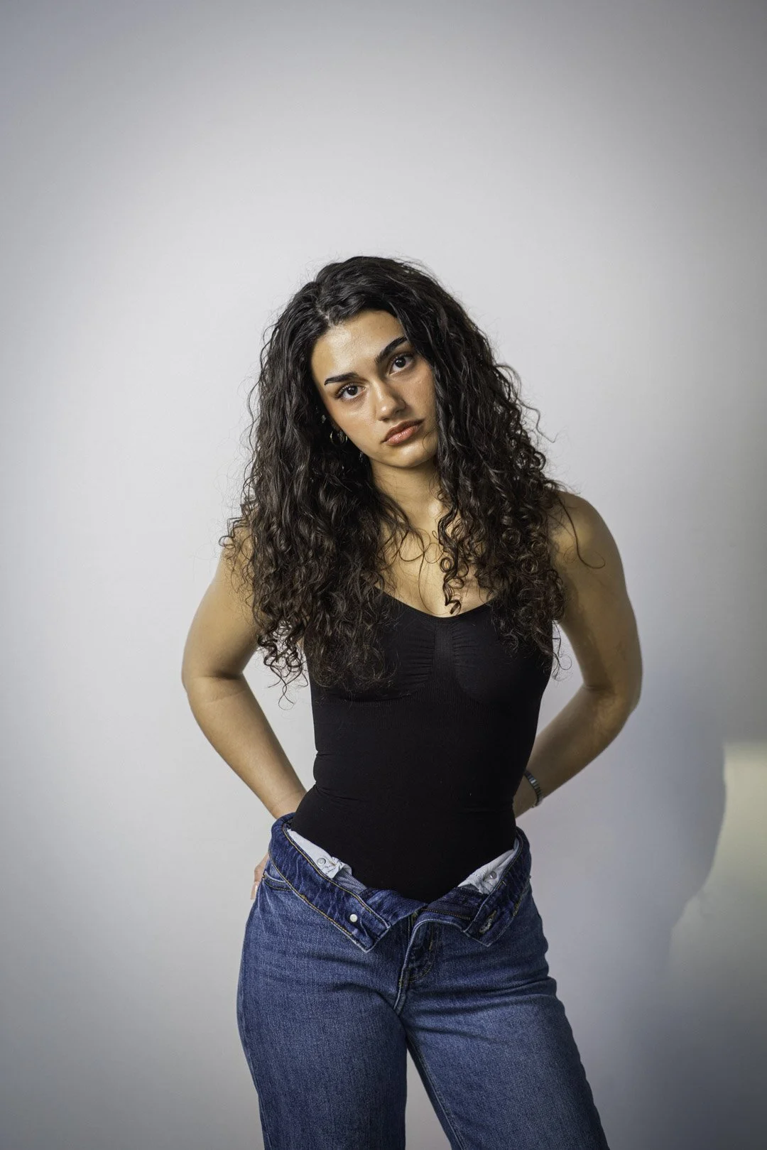 A woman with long, curly dark hair wearing a black sleeveless top and blue jeans, standing against a plain light gray background, looking at the camera with a serious expression.