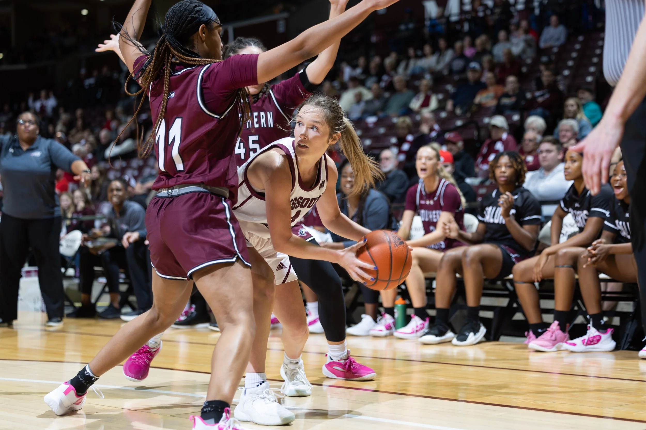A women's basketball game with players on the court, one player in a white jersey holding the ball, defended by a player in a maroon jersey. Spectators sit in the background watching.