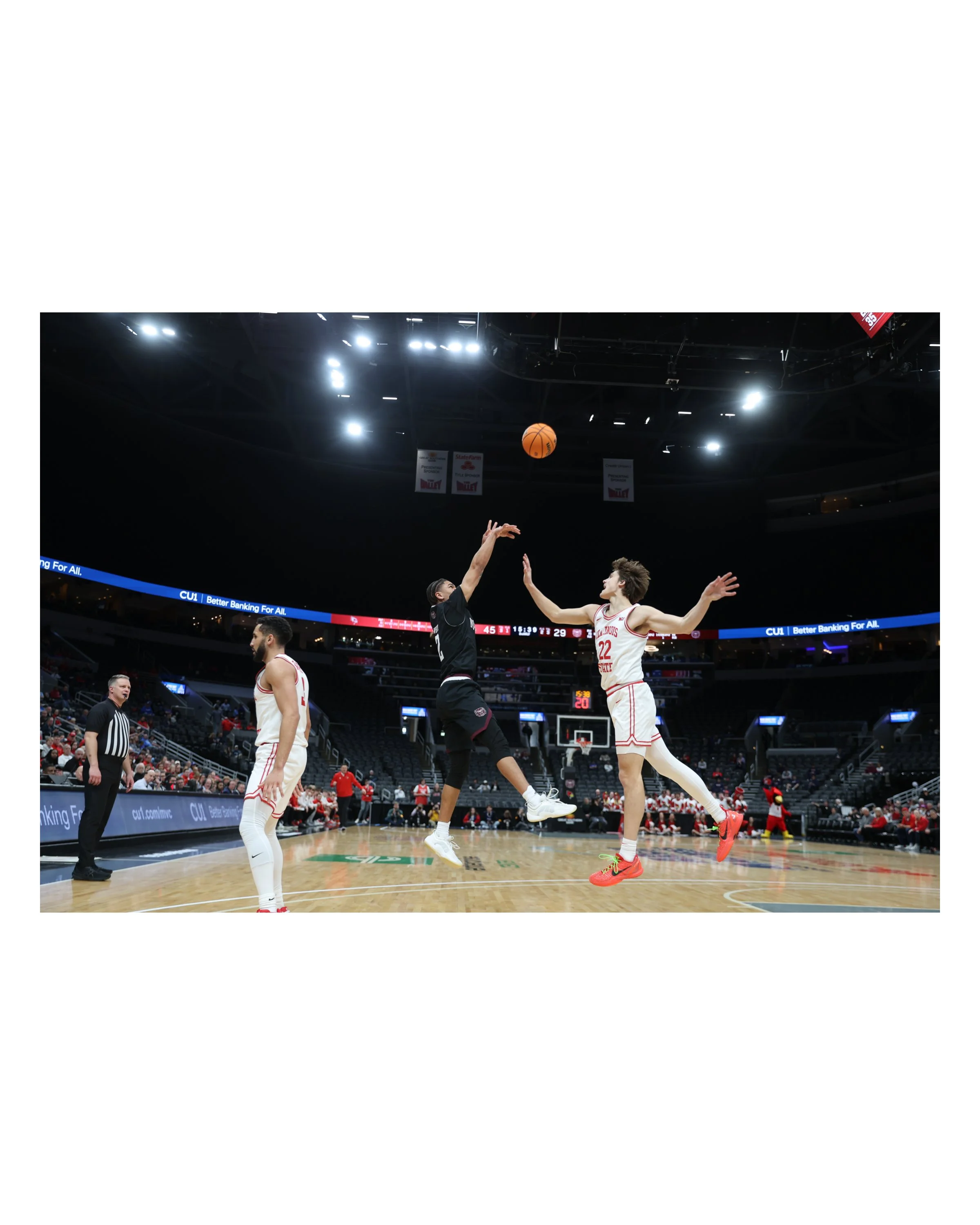 Two basketball players are mid-air reaching for the ball during a game, with another player and referee in the background at an indoor basketball arena.