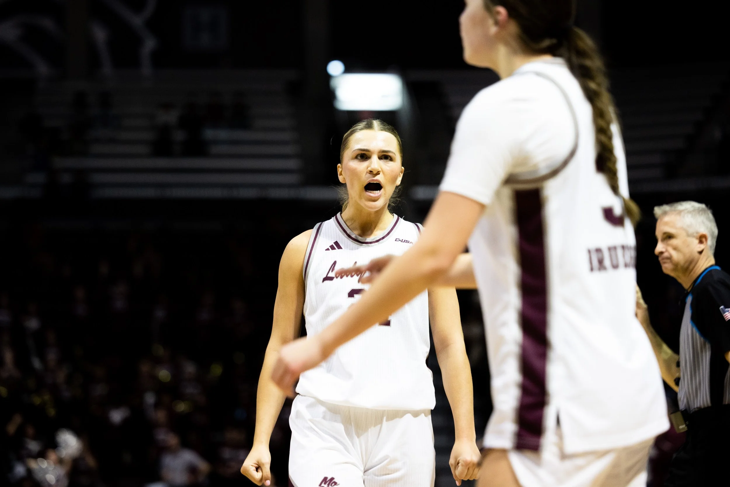 Two female basketball players in white uniforms having an argument on the court during a game, with a referee in the background.