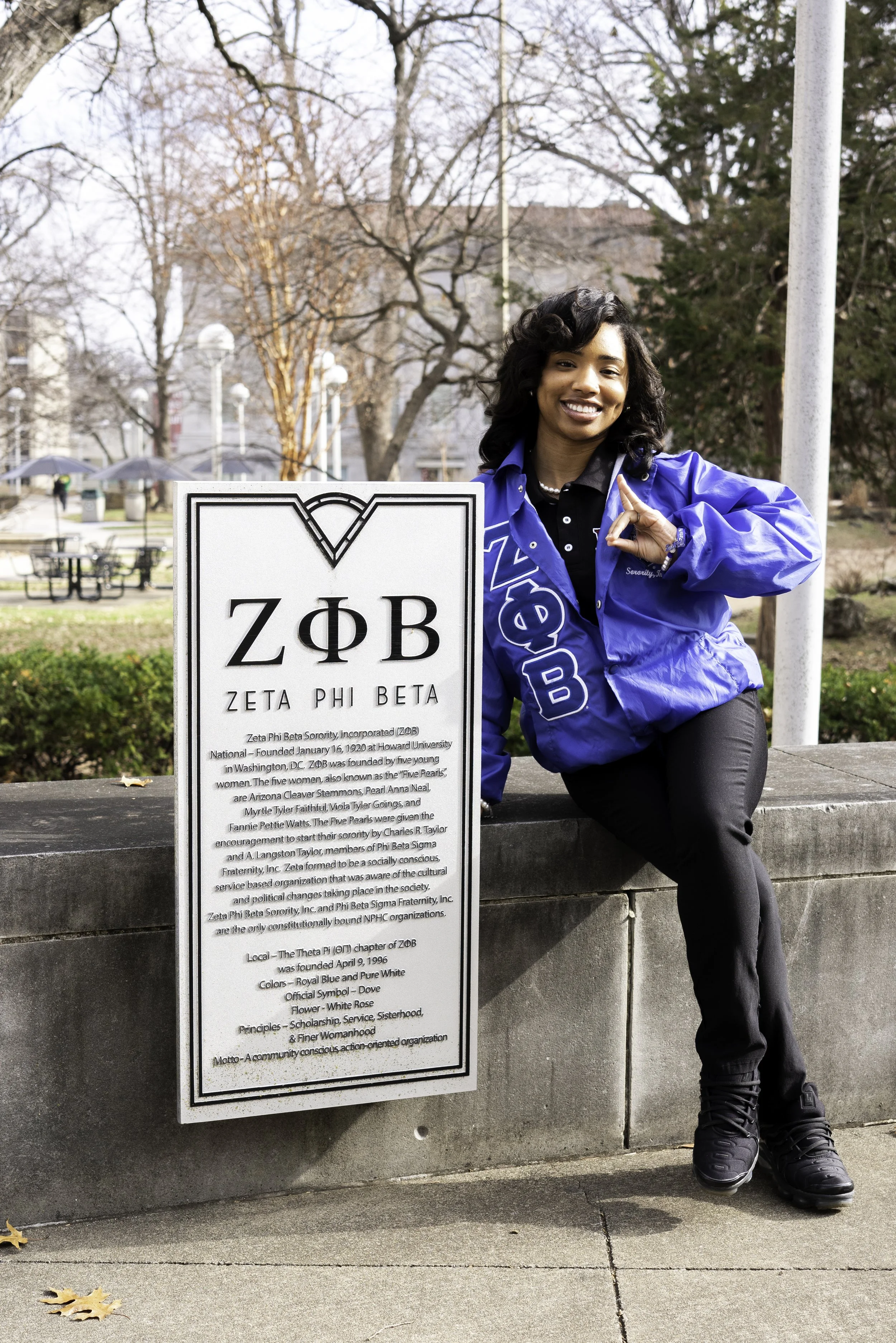 A woman in a blue jacket sitting on a concrete ledge next to a Zeta Phi Beta sorority informational sign in a park with trees and some buildings in the background.