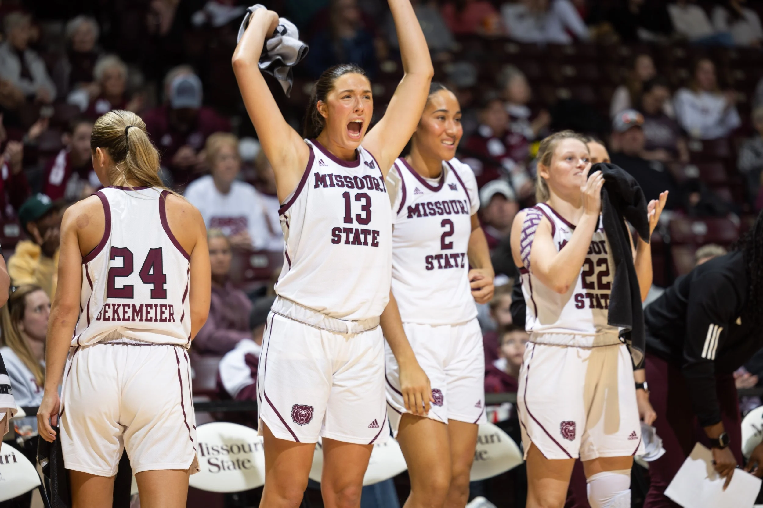 Missouri State women's basketball players on the sideline during a game, with one player cheering and others holding towels and watching.