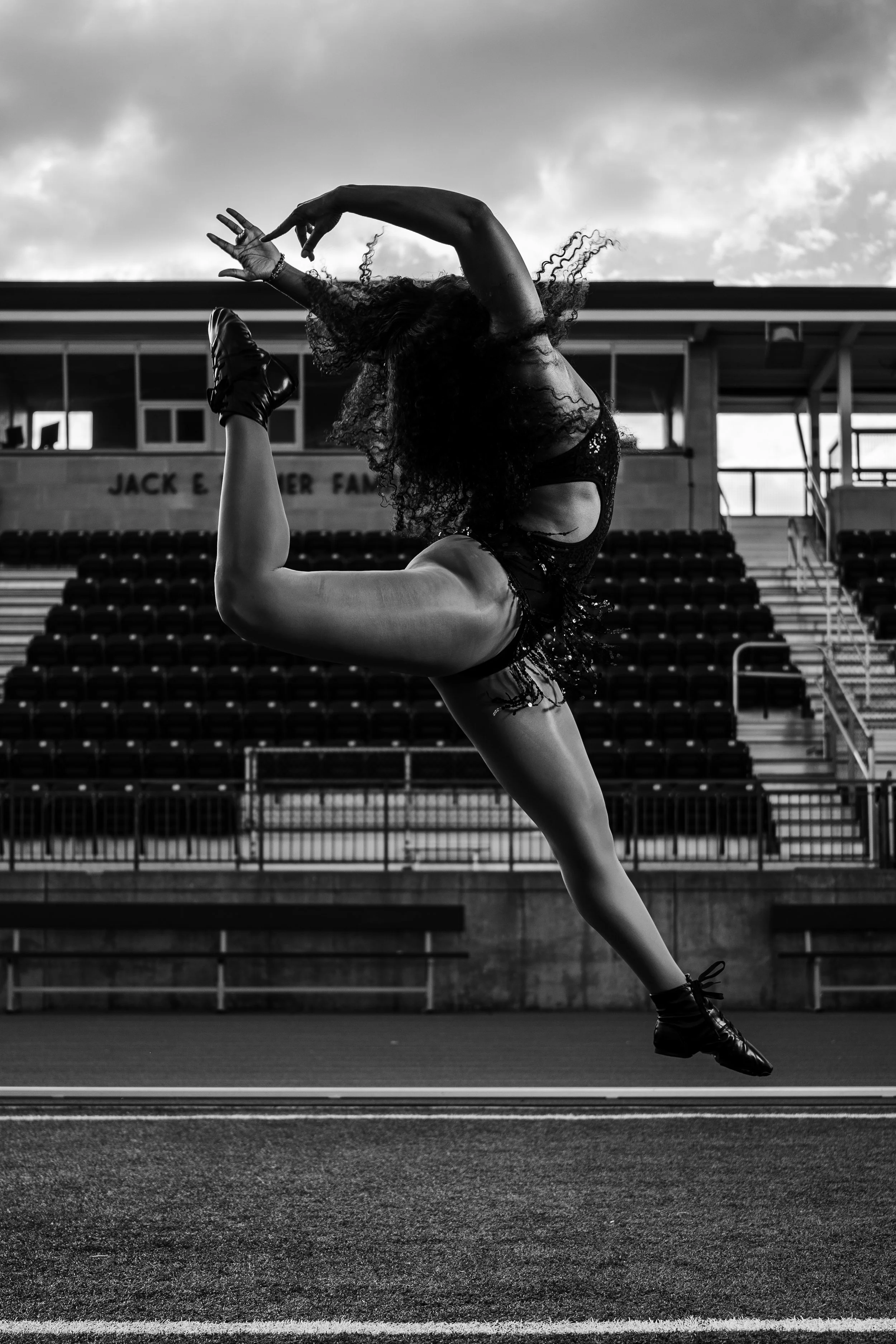 A female dancer in a leotard performs a leap on an outdoor track field, with empty bleachers and a building in the background.