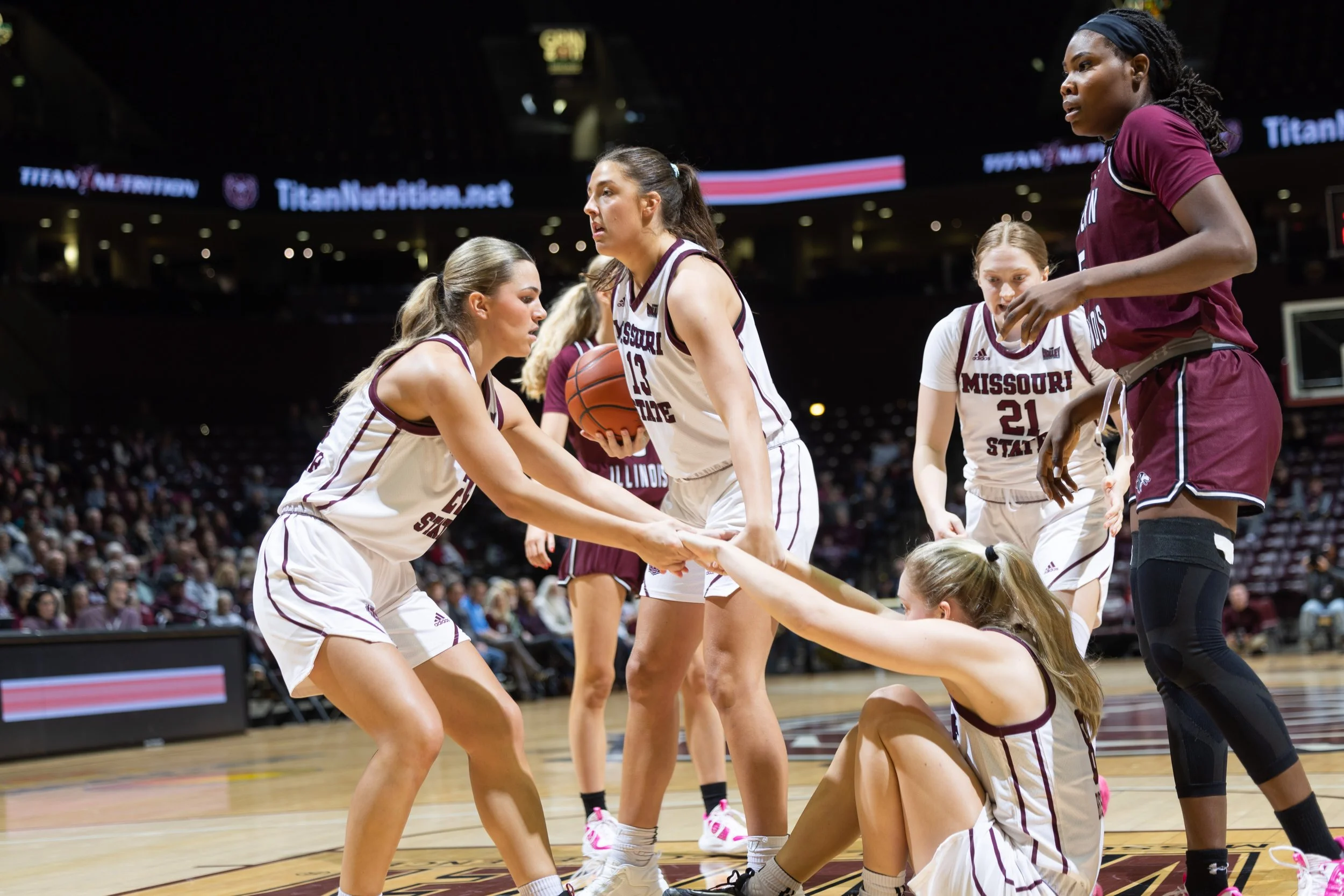 Women basketball players from Missouri State team during a game on the court, with one player on the floor holding hands with a teammate while others look on.