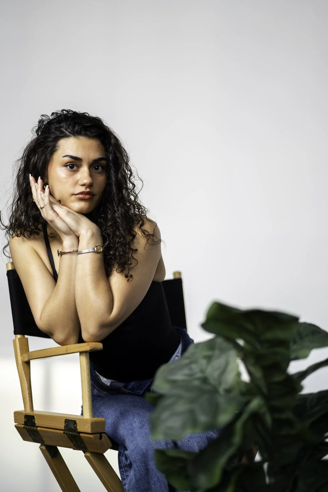 Young woman with curly dark hair, sitting on a wooden director's chair, resting her chin on her hands, looking at the camera against a plain white background, with green plants in the foreground.