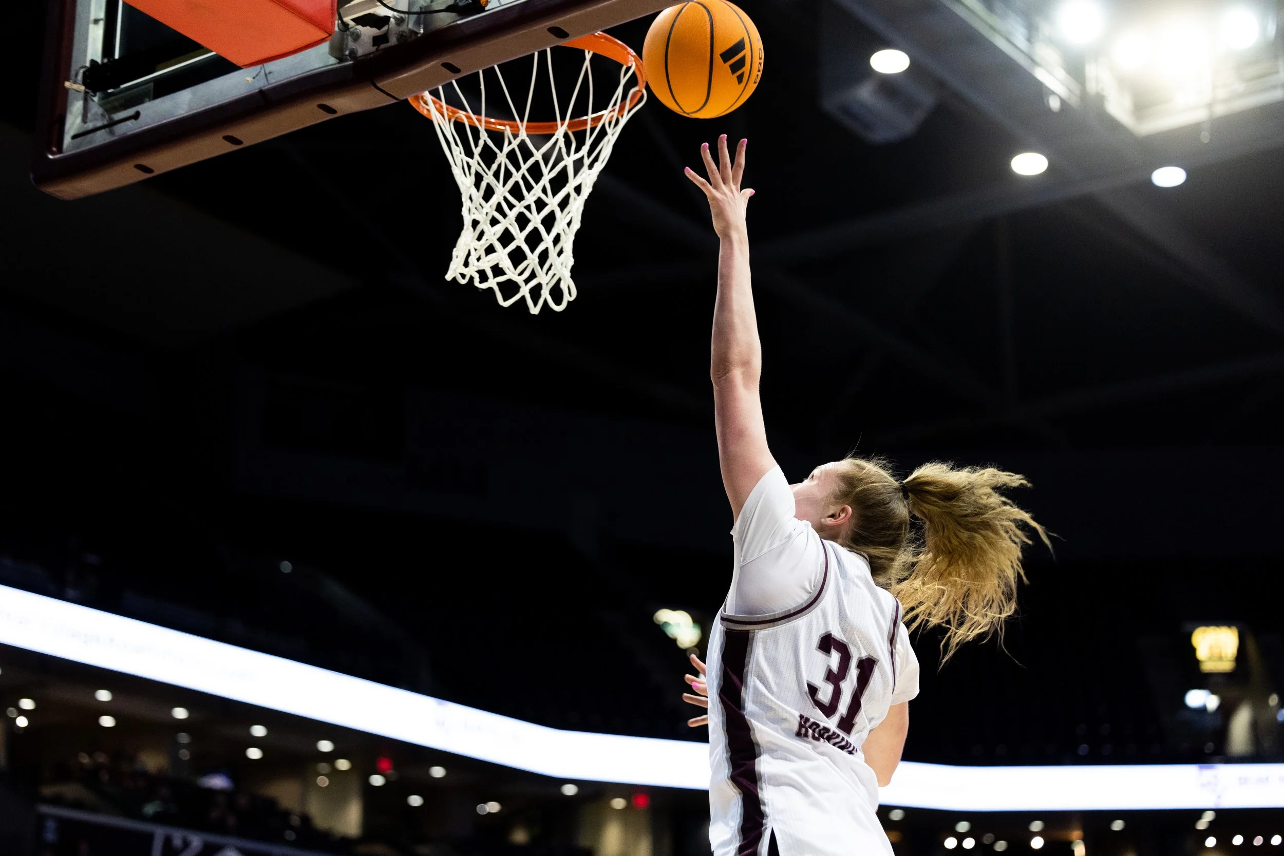 A female basketball player with jersey number 30 jumps to make a shot at the basket, with the ball approaching the hoop, in an indoor basketball arena.