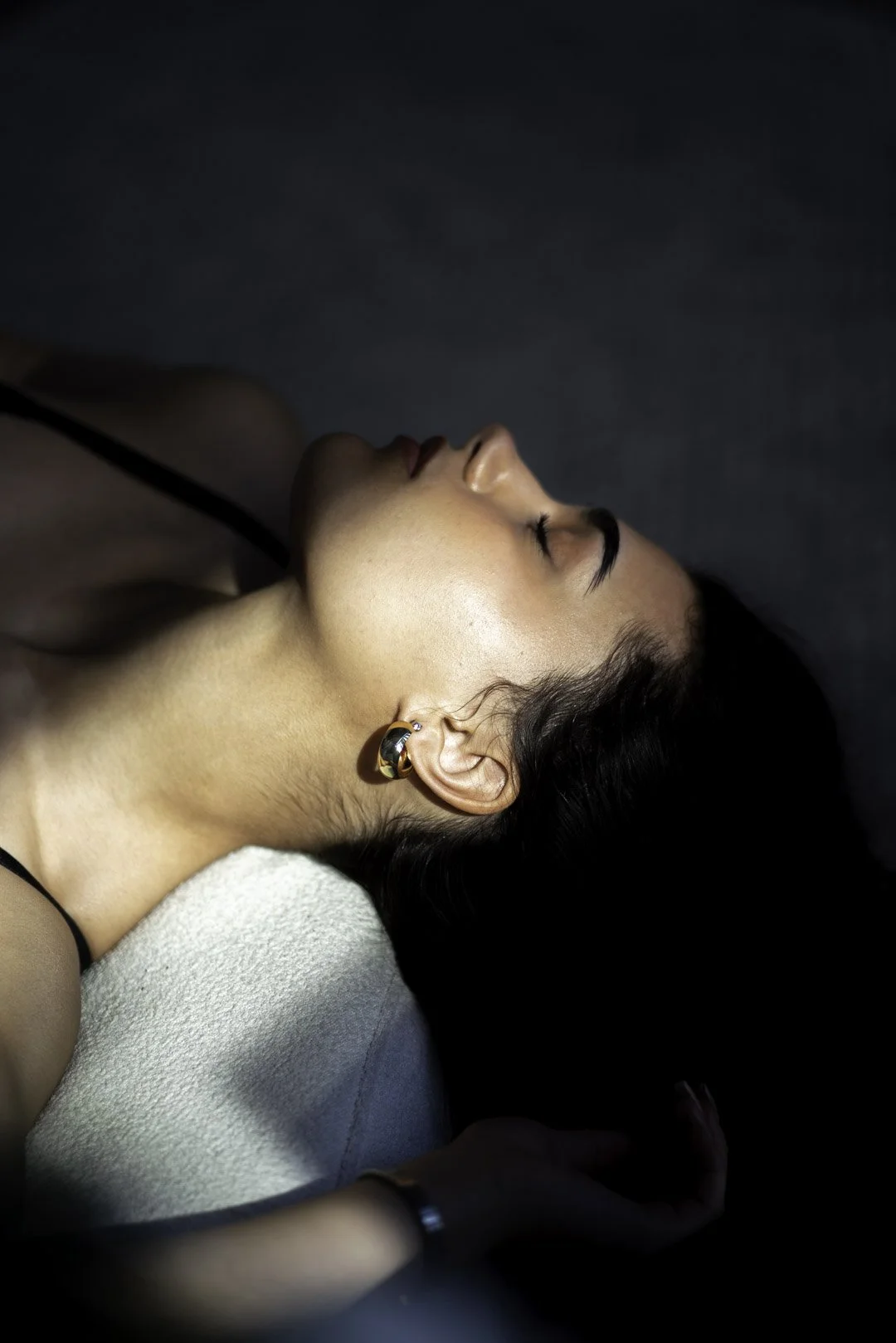 A woman with dark hair lying on her side with her eyes closed, wearing a black spaghetti strap top and a hoop earring, on a white pillow against a dark background.