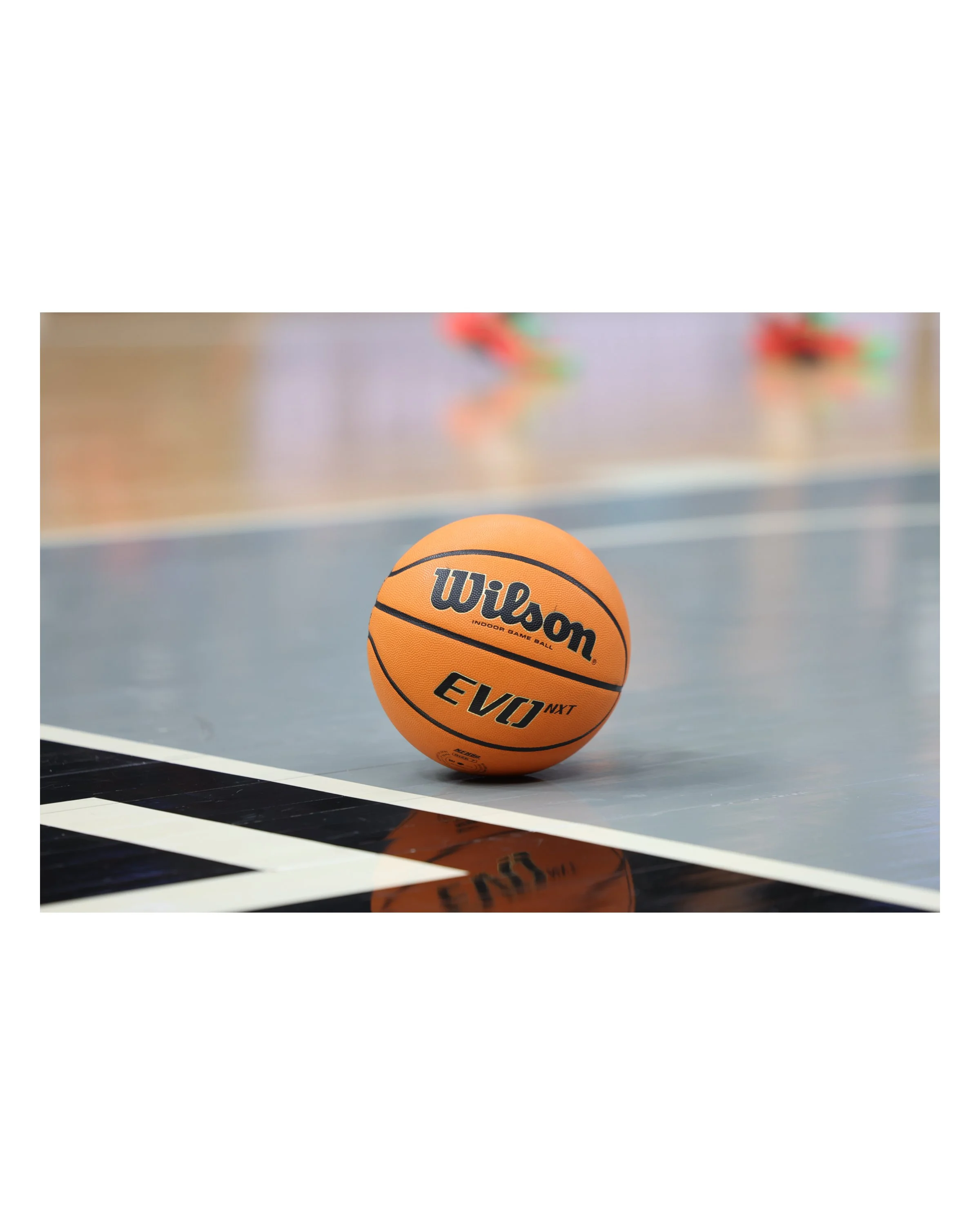 An orange Wilson basketball resting on a gray and black indoor basketball court with a blurred background of players.
