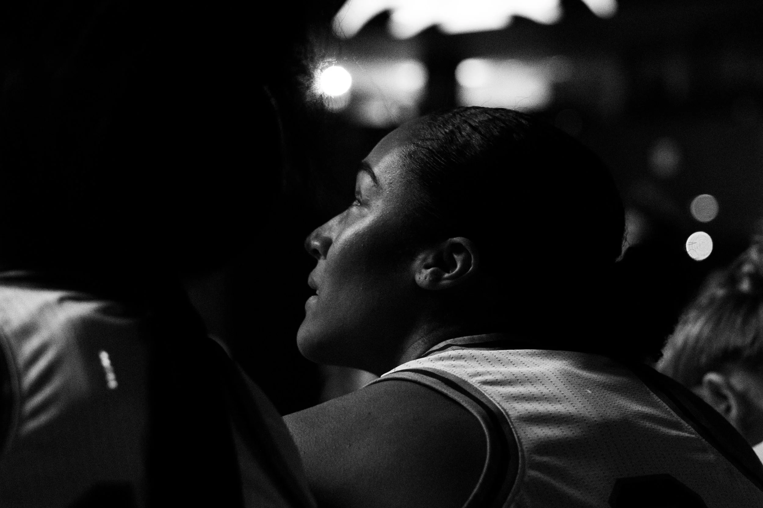 A black-and-white photo of a woman with slicked-back hair looking upward, illuminated softly from the side, with other people around her in a dimly lit environment.