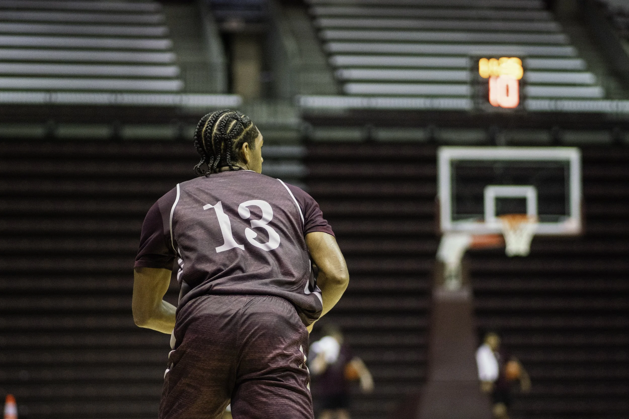 A person with braided hair wearing a maroon sports jersey with the number 13 on the back, standing on a basketball court, with a basketball hoop in the background and a shot clock displaying 8:45.