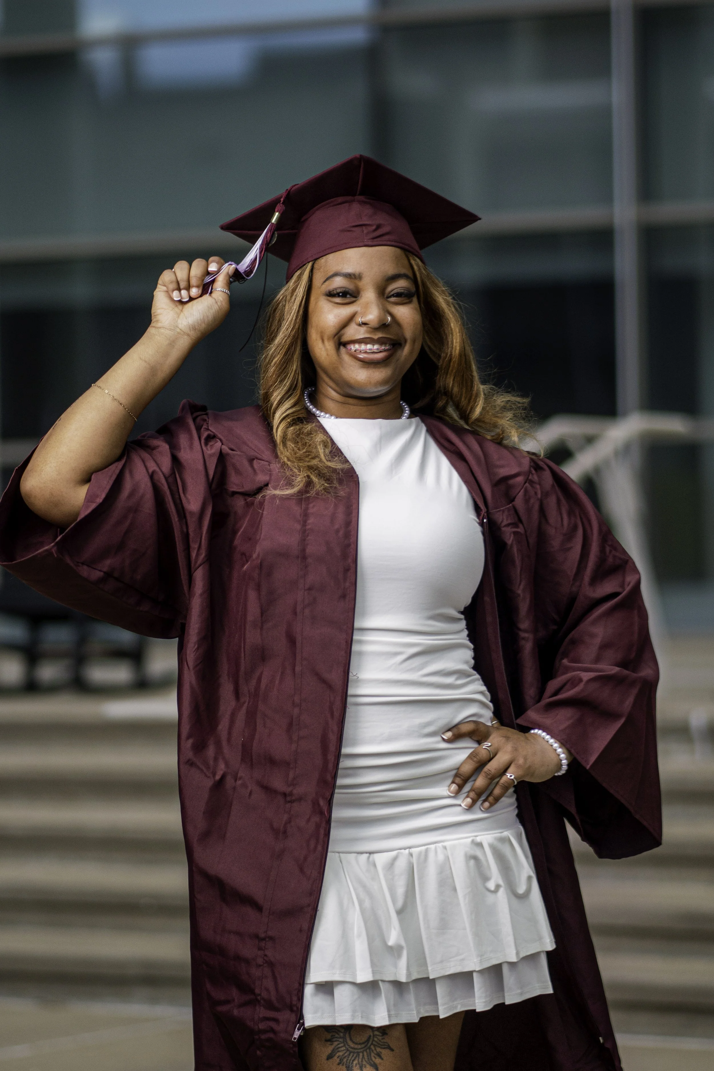 A woman in graduation cap and gown smiling, holding her tassel, standing outdoors with modern glass building background.