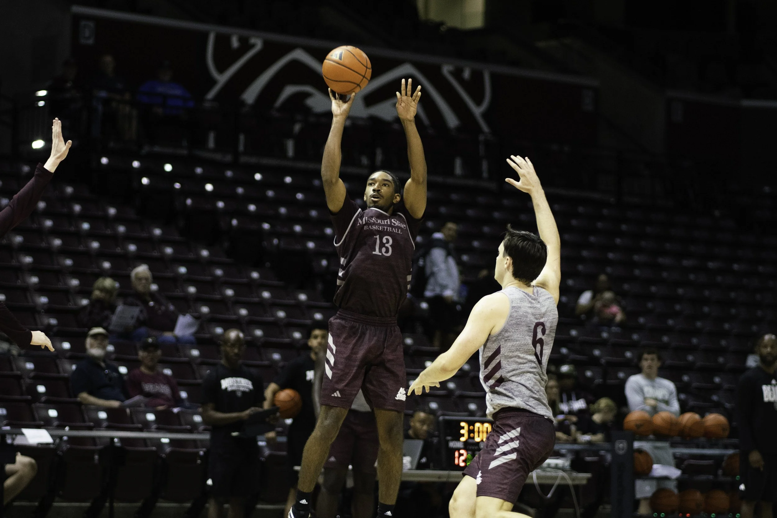 Two basketball players in maroon and gray uniforms compete on the court. One player is jumping to shoot or pass the ball, while the other player is defending with an arm raised. Spectators watch from the stands in the background.
