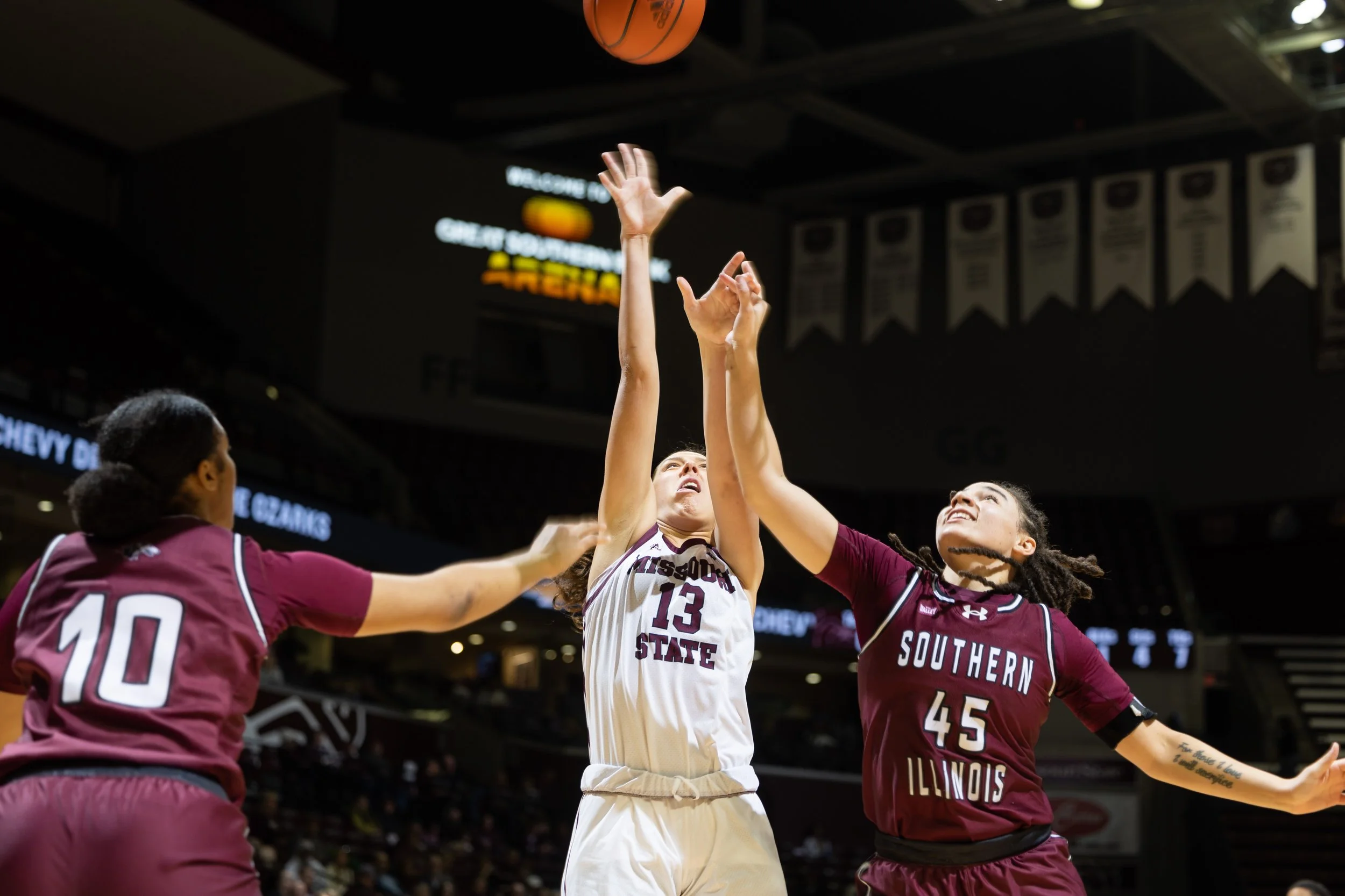 A women's basketball game with three players reaching for the ball, with one player from Missouri State in a white uniform jumping and two players from Southern Illinois in maroon uniforms attempting to block or catch the ball.