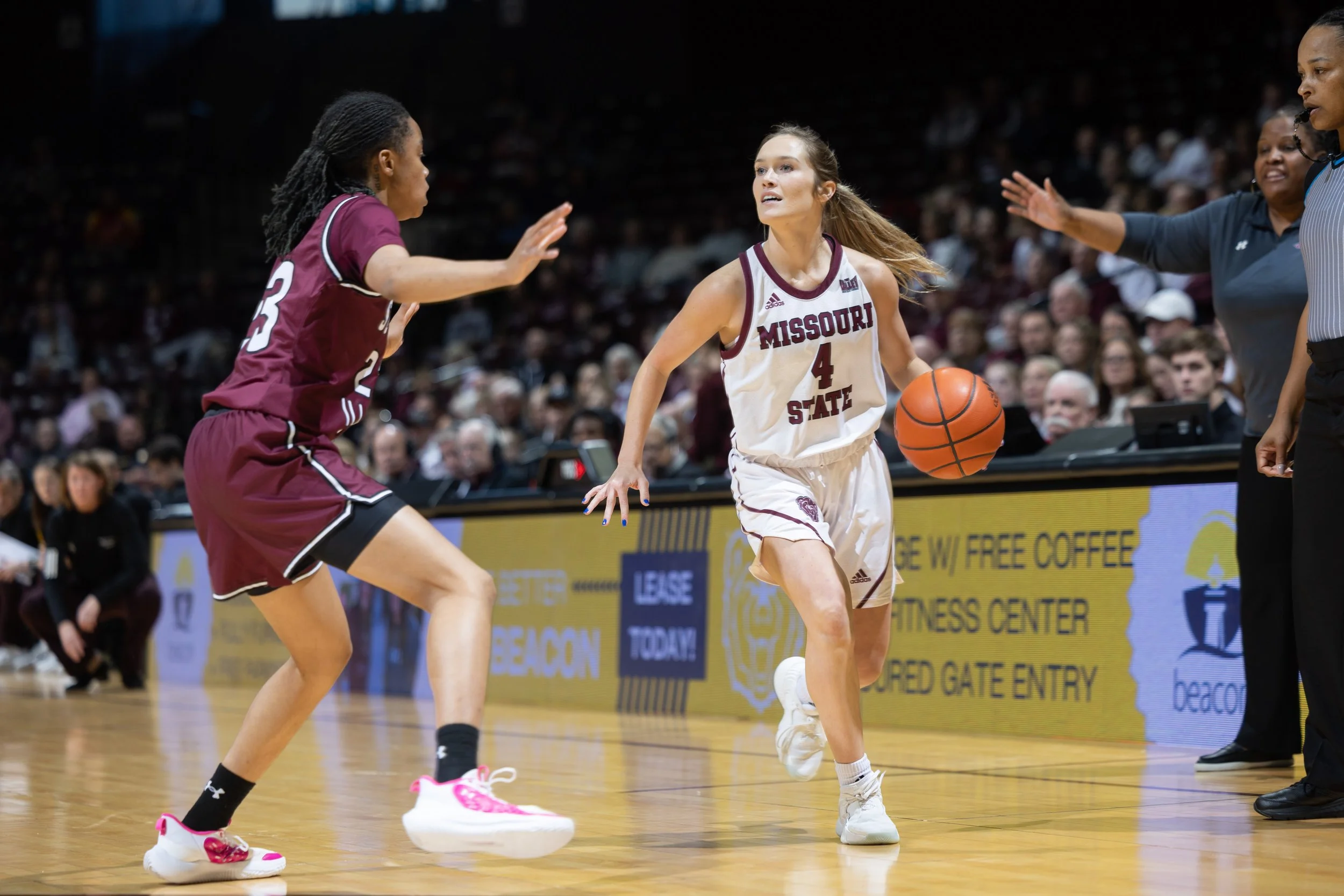 A women's basketball game with one player from Missouri State driving towards the basket, guarded by a player from an opposing team, while an referee observes.