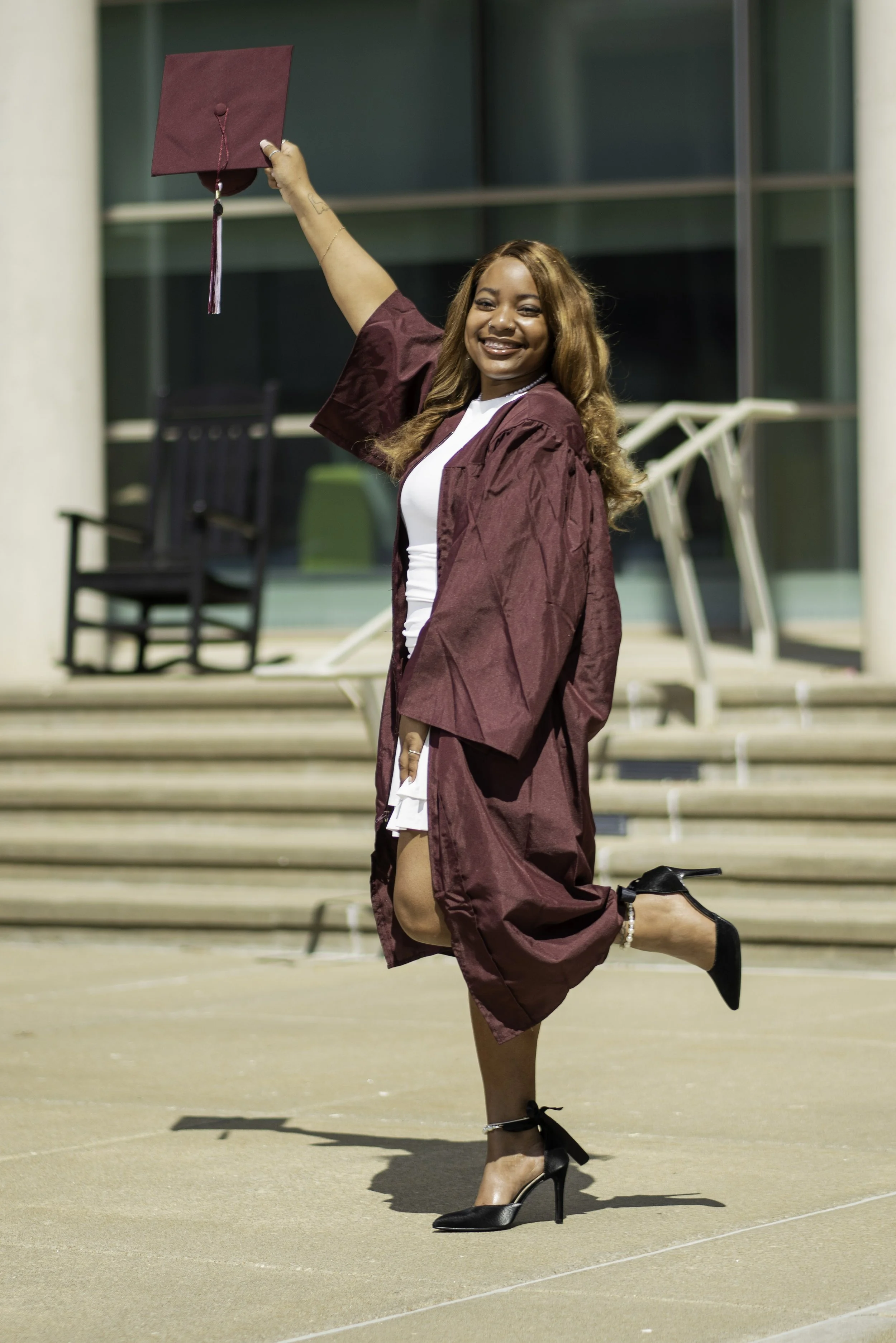 A young woman in a graduation cap and gown celebrating outdoors, holding her cap in the air and smiling.