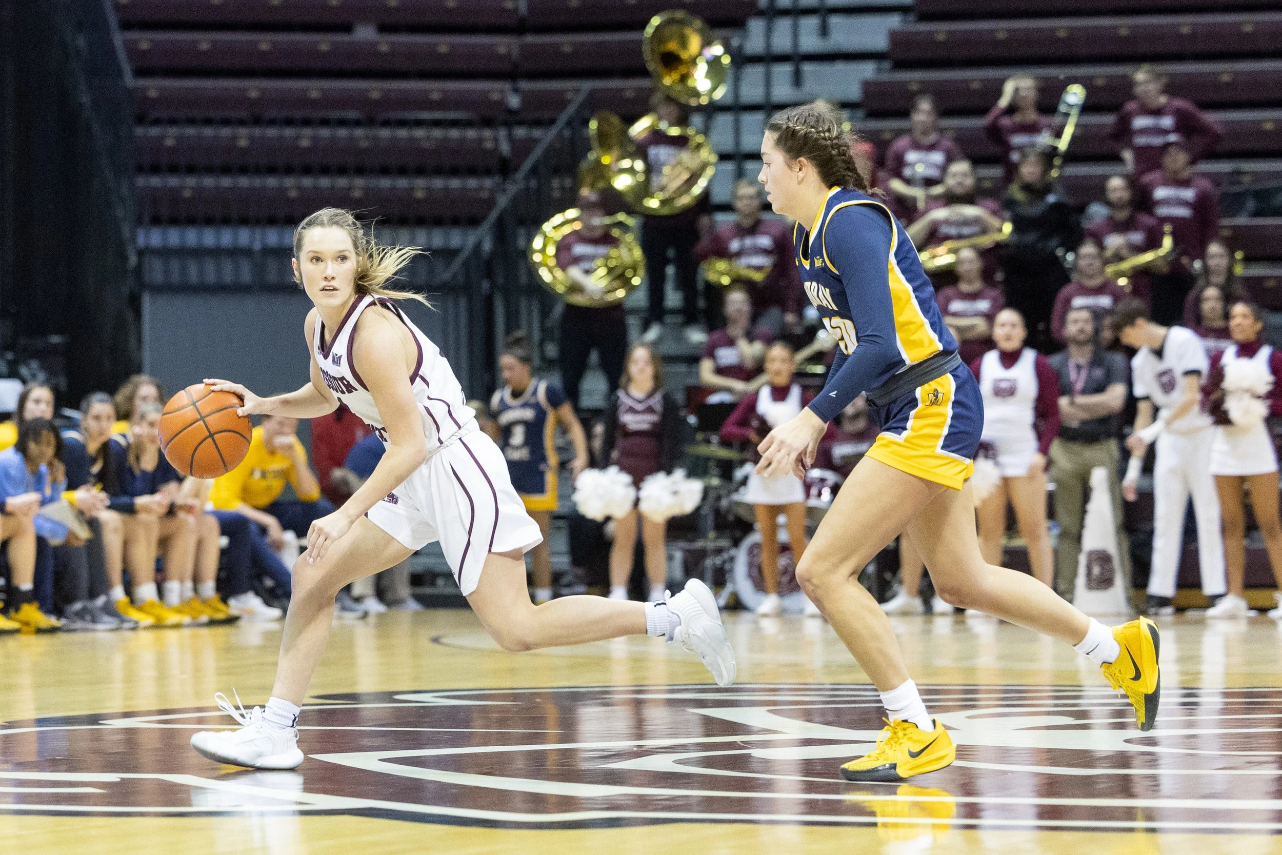 Two female basketball players in action during a game, with one dribbling the ball and the other defending, on an indoor basketball court with spectators and a marching band in the background.