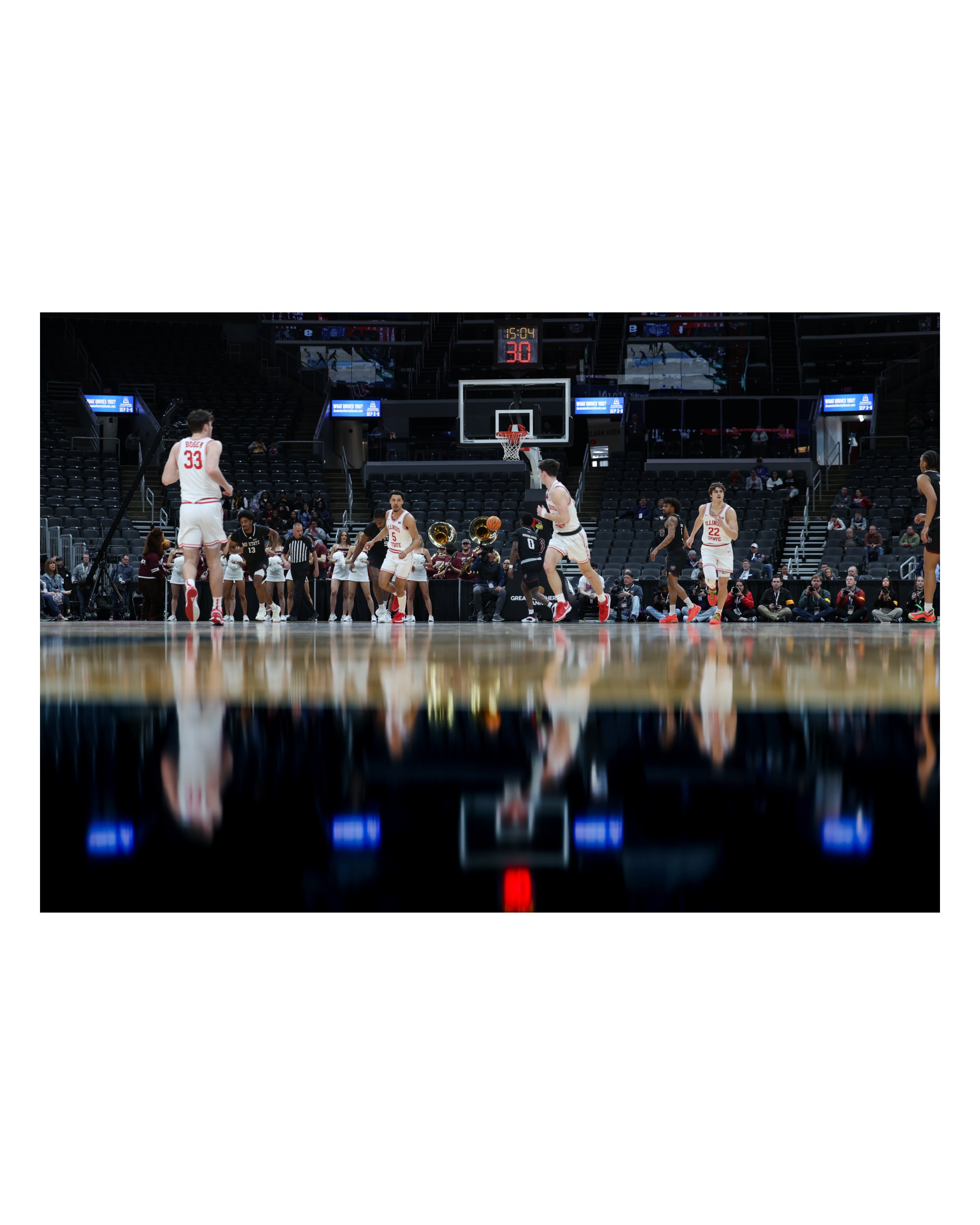 A basketball game in progress with players on the court, cheerleaders, and a band in the background, in a large indoor arena.