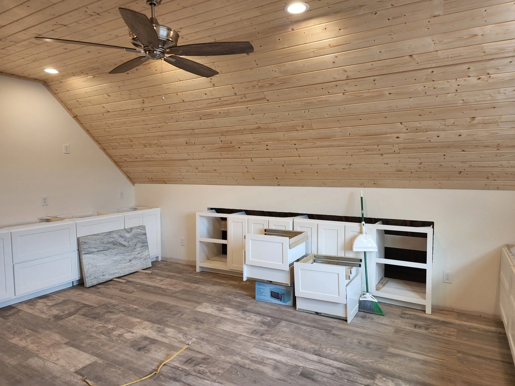 Room under construction with kitchen cabinets, a marble backsplash, and a wooden ceiling and floor. Construction materials and tools are visible, including boxes, a broom, and a yellow extension cord.