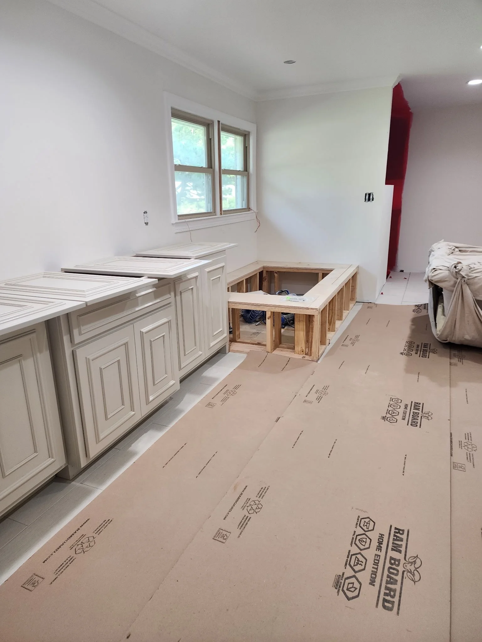 Partially completed kitchen renovation with white cabinets, a window, and flooring in progress, with protective sheeting on the floor.