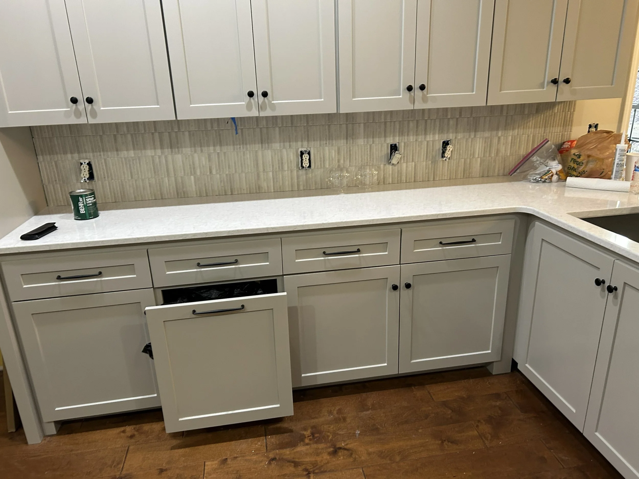 Kitchen with white cabinets, beige tiled backsplash, and various items like a can, plastic bags, and a roll of paper towels on the countertop.