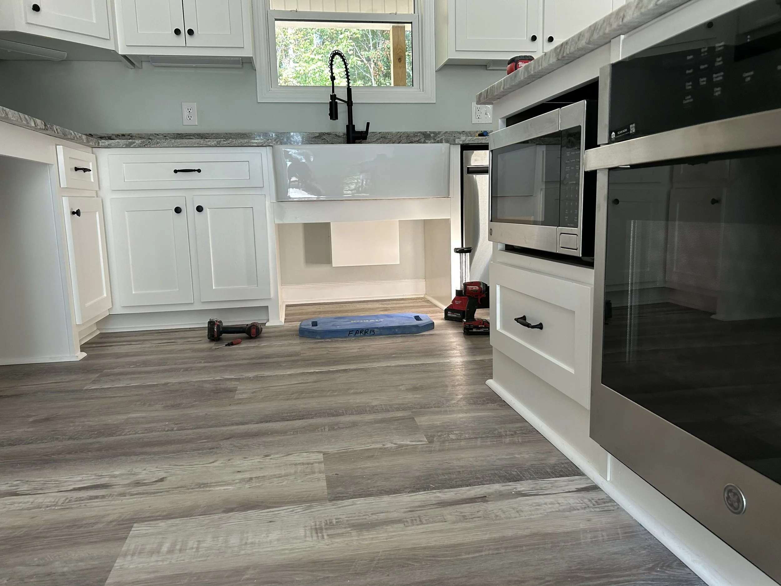 Kitchen with white cabinets, a window above the sink, a black faucet, and a gray marble countertop. There are tools on the floor, including a drill, a red cordless tool, a blue Farris step, and a red drill on a stand.
