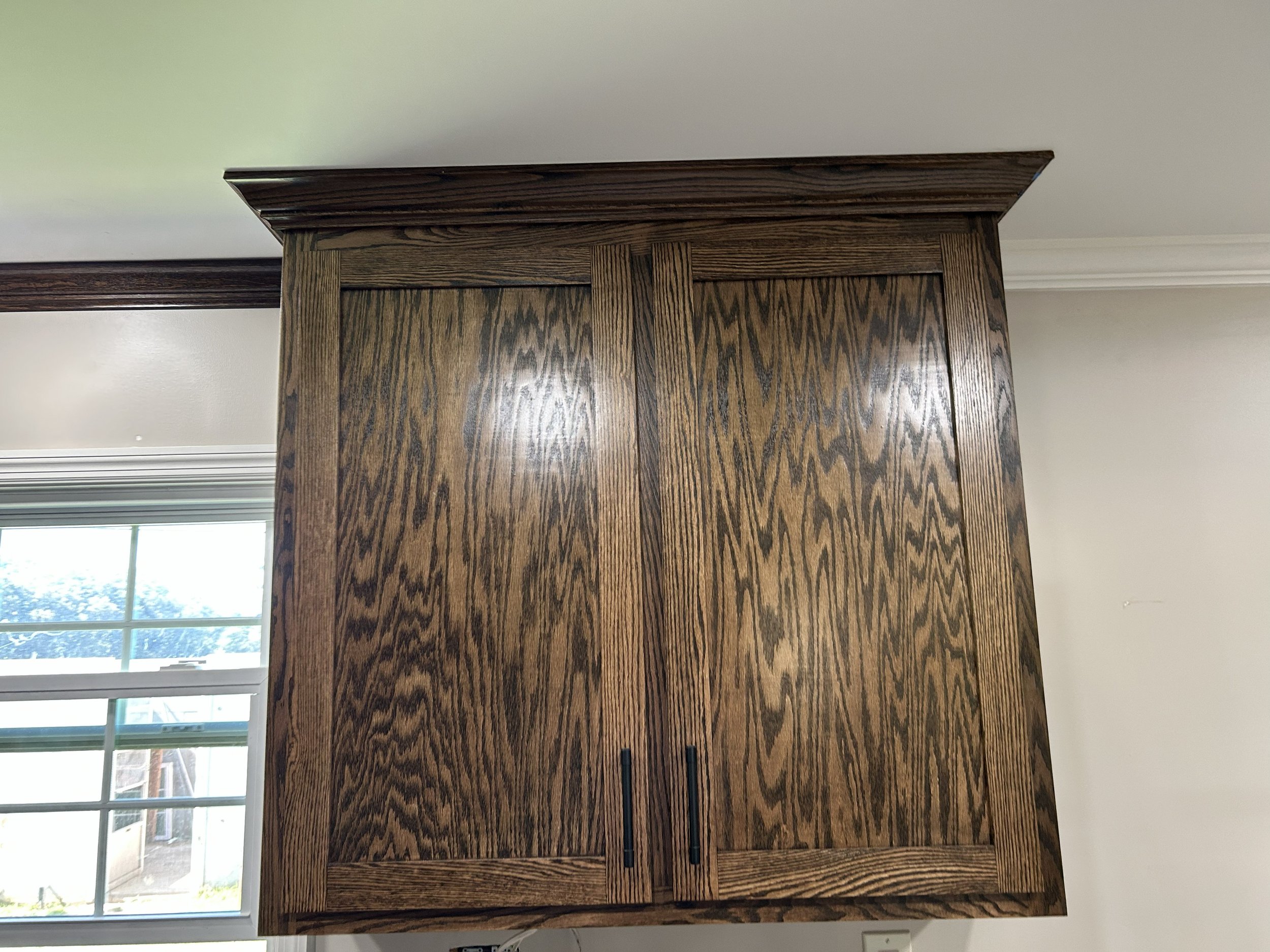 Wooden kitchen cabinet with two doors and black handles, mounted on white wall near window.