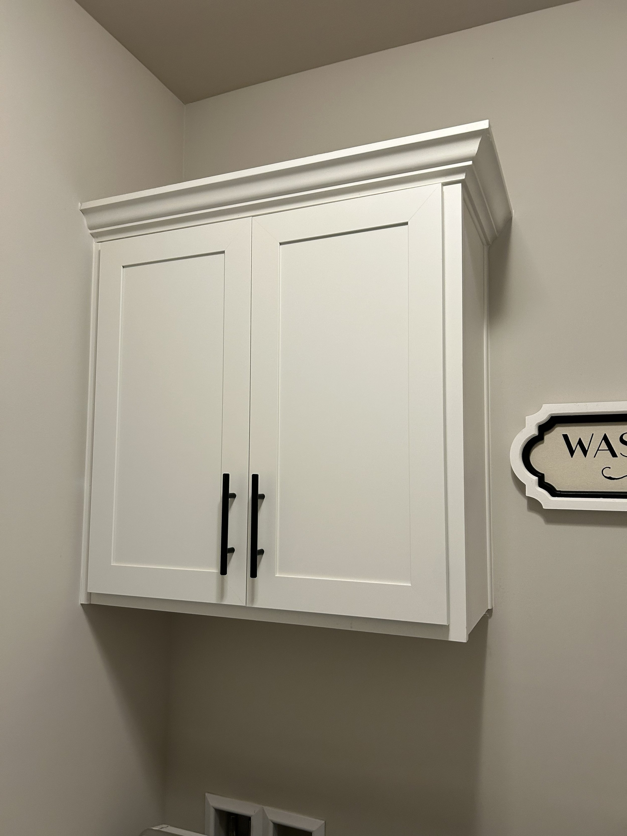 White wall-mounted cabinet with black handles, decorative crown molding, mounted on a beige wall in a room.