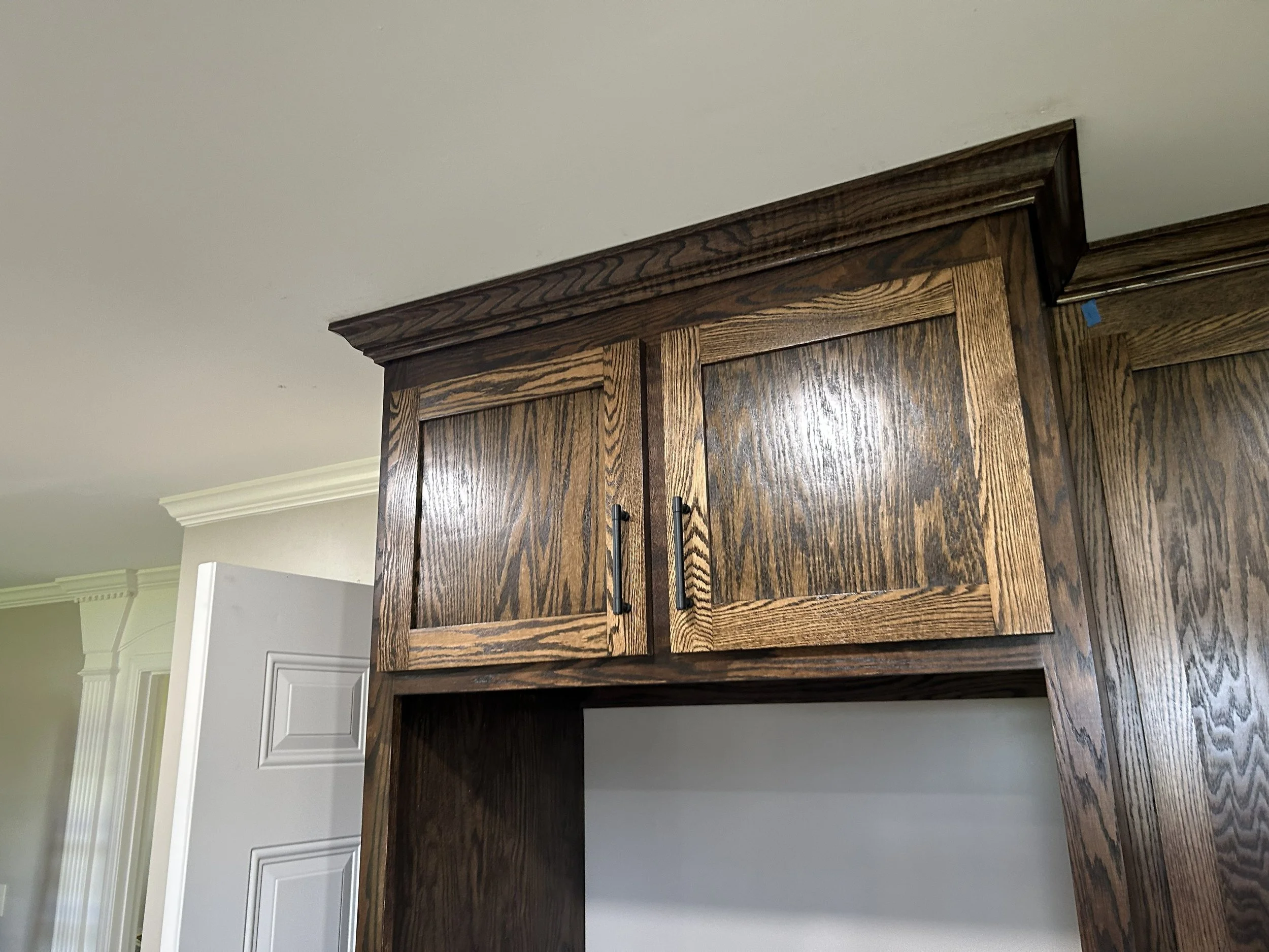 Close-up of a wooden kitchen cabinet with two doors, showing grain patterns and black handles, mounted on a wall with a white door and crown molding in the background.