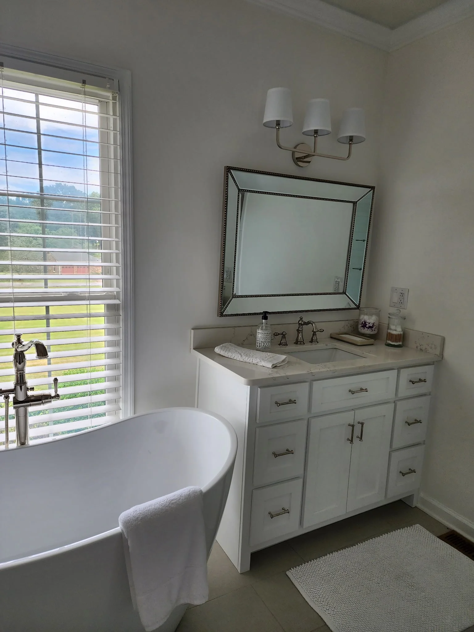 A bathroom with a white vanity, a large mirror with a decorative frame, a bathtub, and a window with white blinds showing a landscape view. Decor includes a candle jar, a soap dish, and a small jar with brushes, with a white rug on the gray tile floo