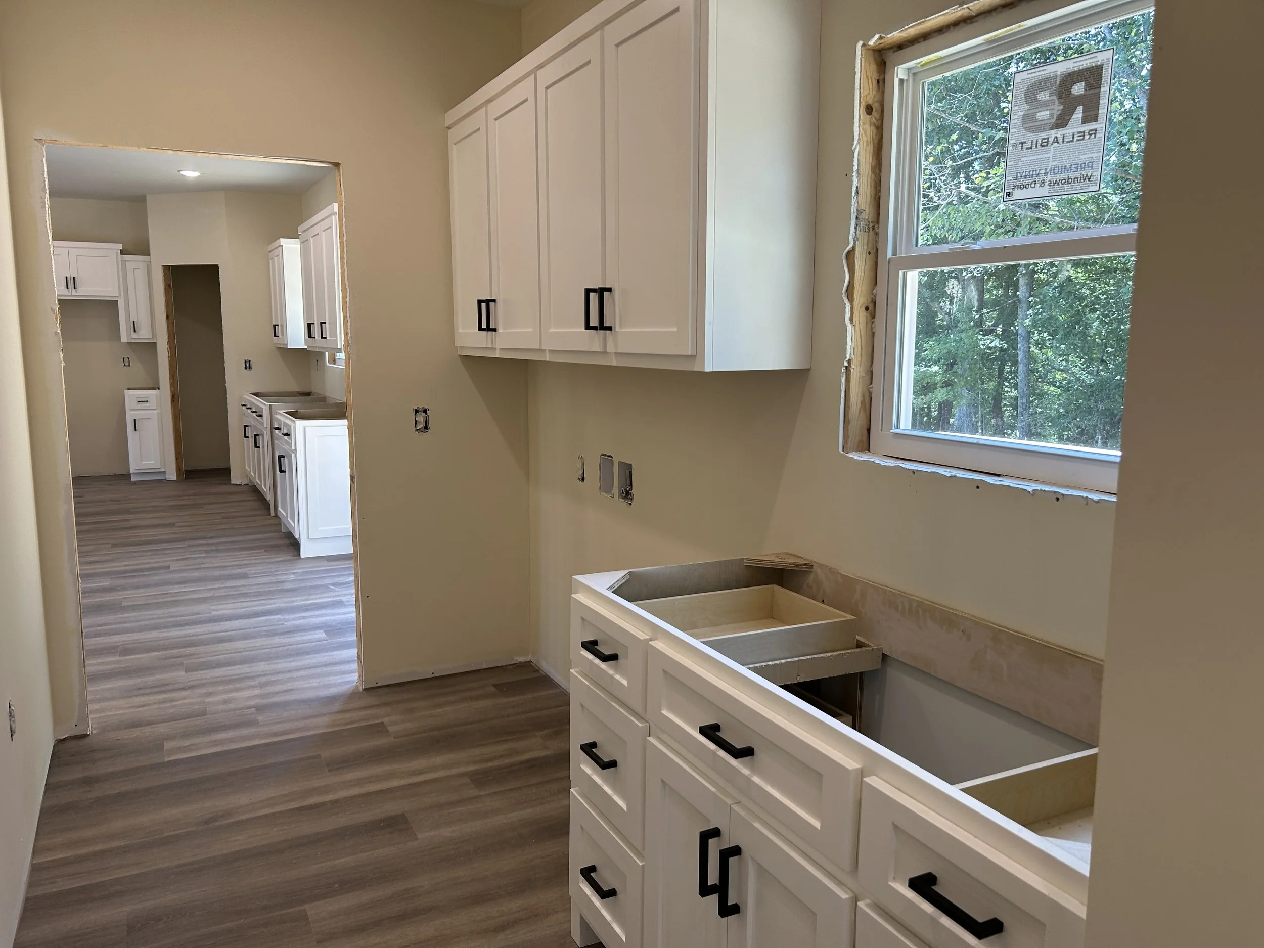 Kitchen under renovation with white cabinets, wood flooring, and a window with a view of trees, showing unfinished window trim and wiring outlets.