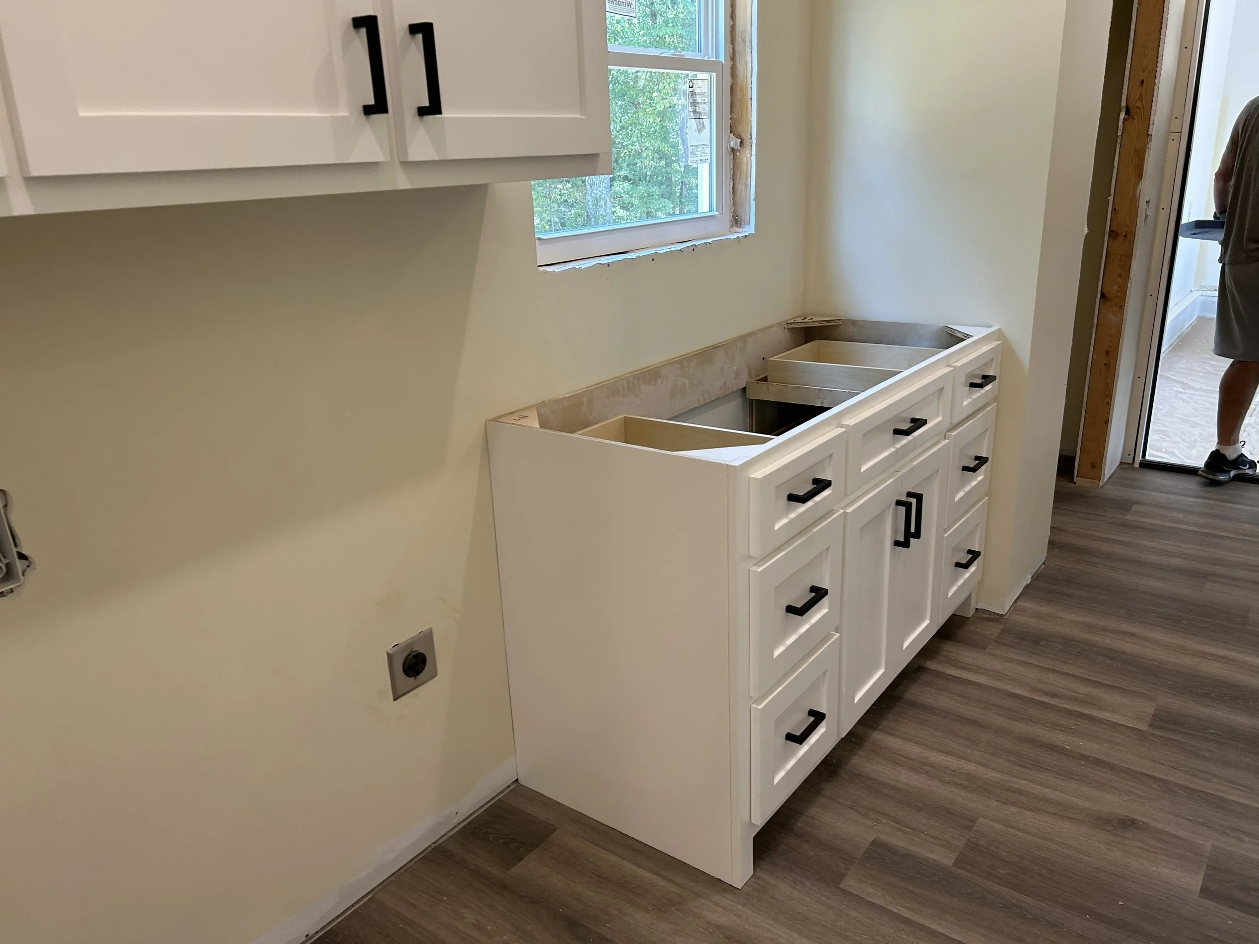 Kitchen cabinet installation in progress, with white cabinets and black handles, and a window above.