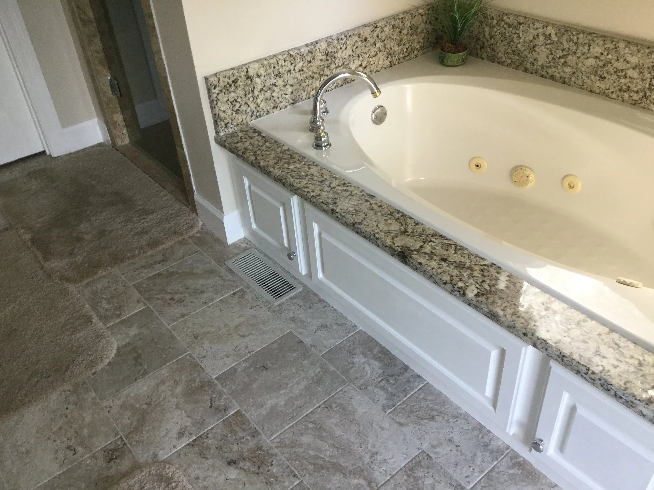 Bathroom featuring a granite-topped bathtub with jets, a silver faucet, white cabinet panels below, a small green plant on the ledge, beige tiled floor, an air vent, and a bathtub with jets.