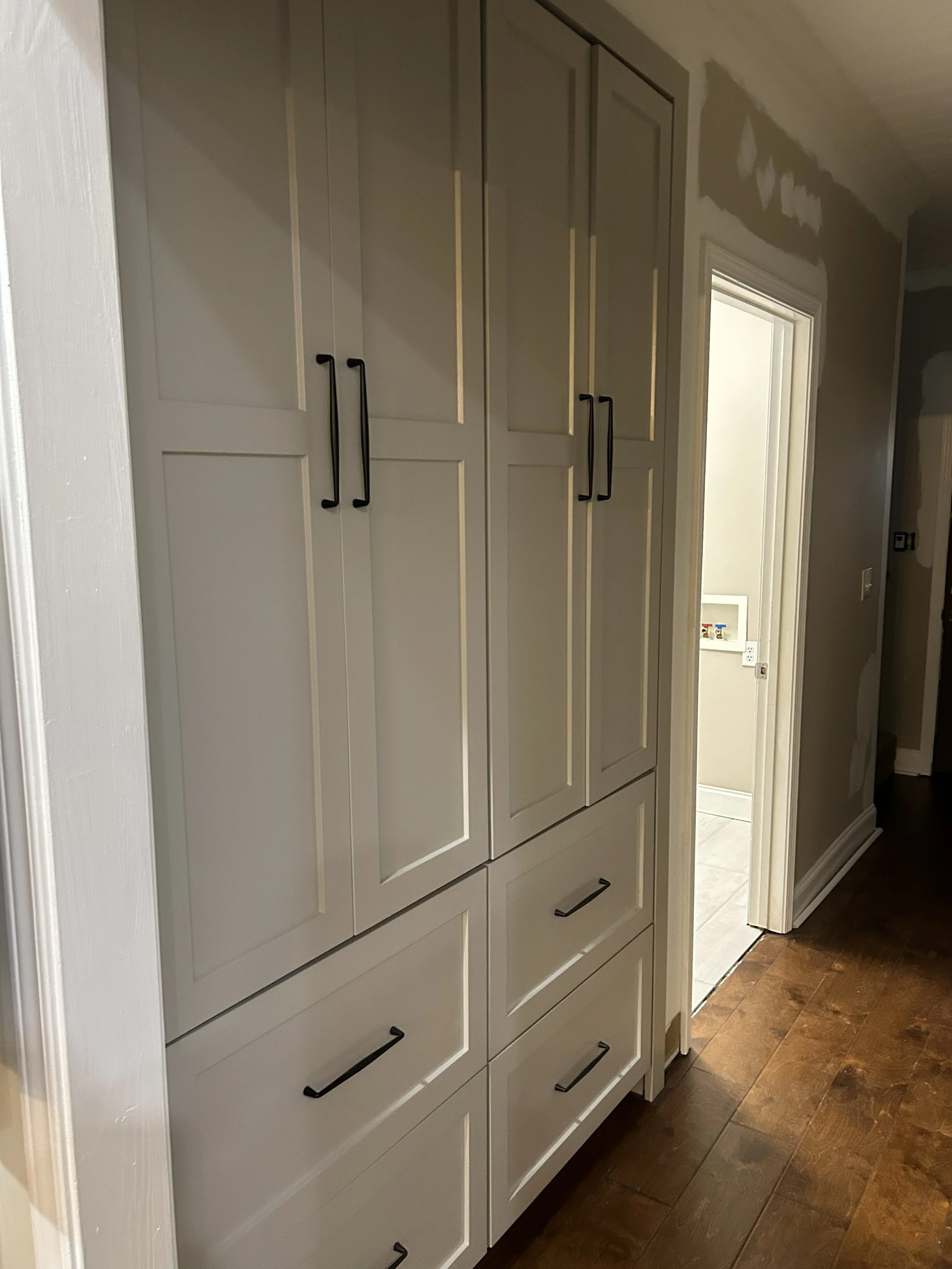 White kitchen cabinet with black handles and wood floor, doorway leading to a laundry area with visible water hookups.