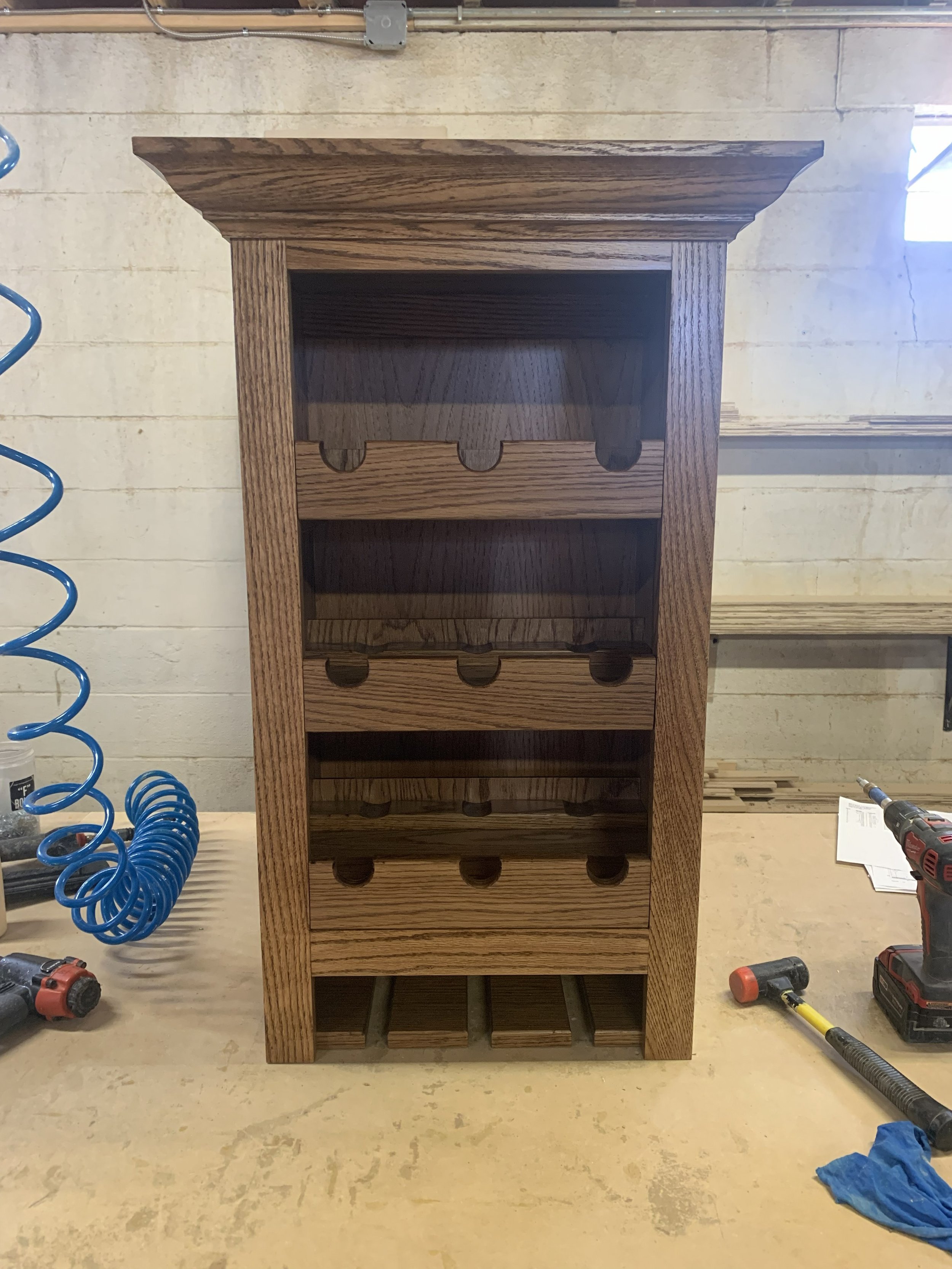 A handcrafted wooden wine rack with four shelves and a slatted base, made of dark wood, placed on a workbench in a workshop.