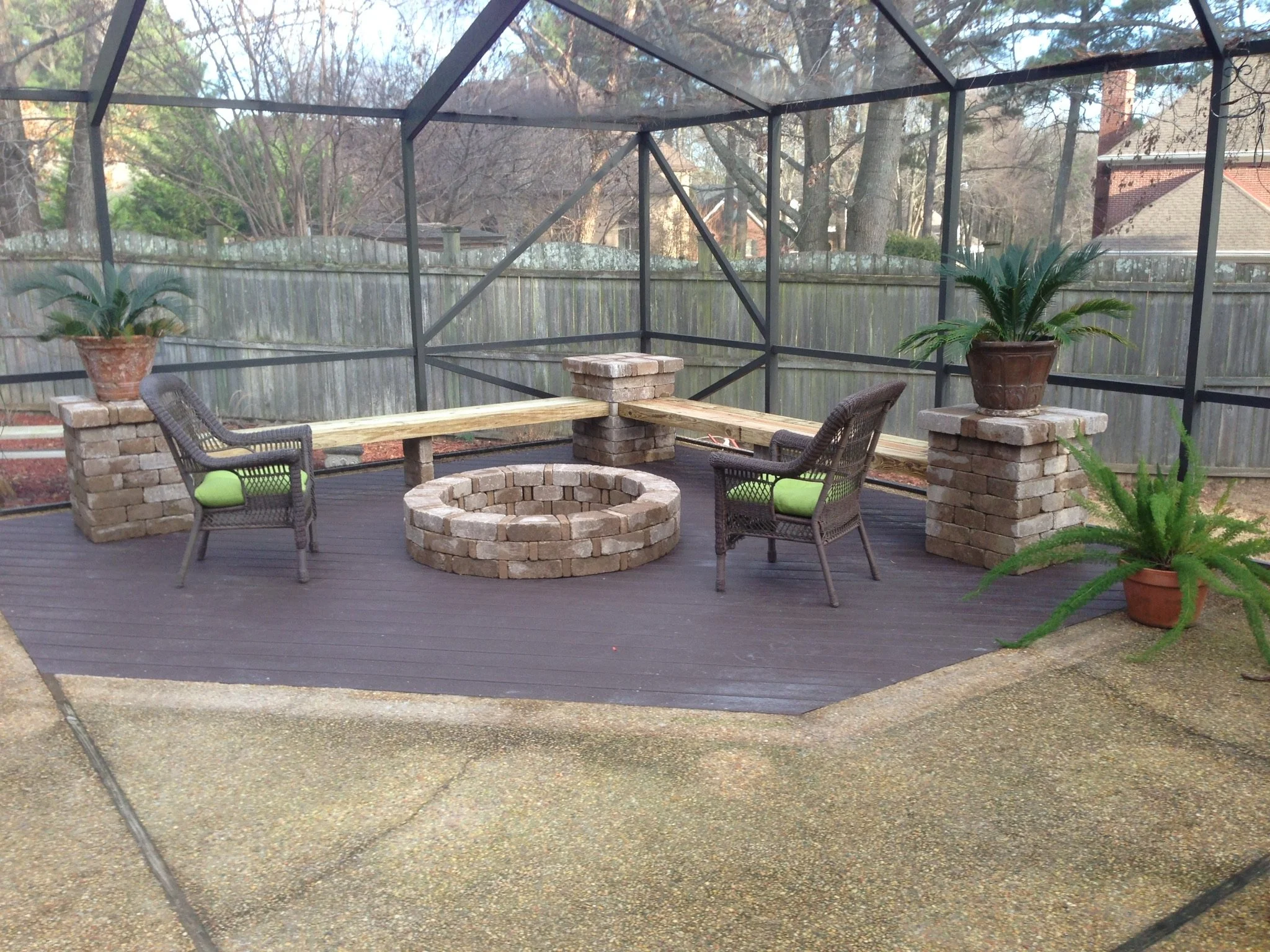 A backyard patio with a screened enclosure, two potted plants on brick pillars, two wicker chairs with green cushions, a circular brick fire pit, and a wooden bench on brick supports.