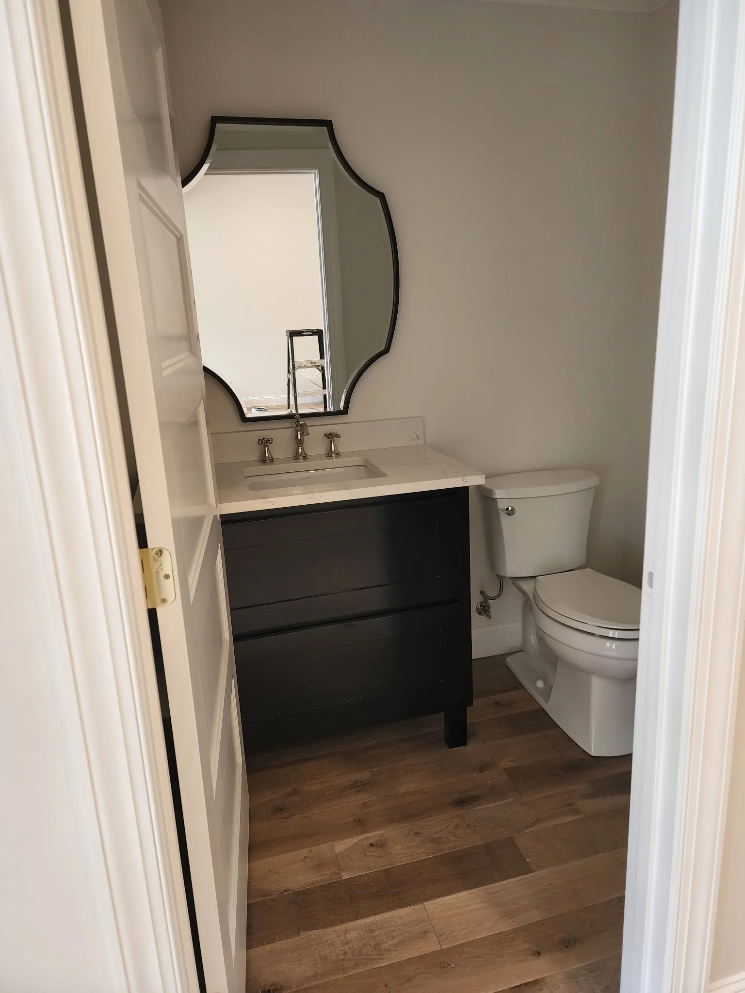 Small bathroom with wooden flooring, a black vanity with a white marble countertop, a modern mirror with a black frame, a sink, a double-handle faucet, and a white toilet.