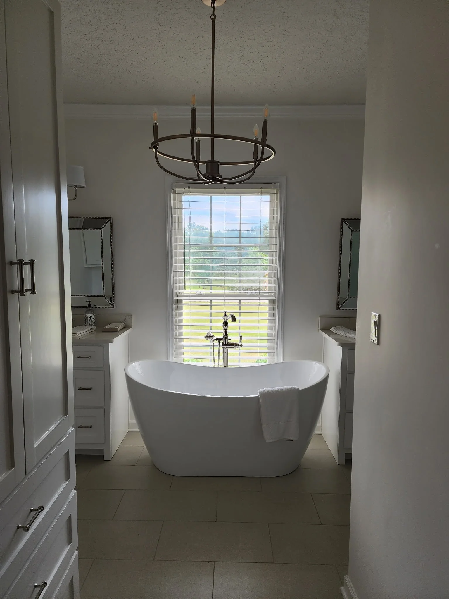 A modern bathroom with a freestanding bathtub in front of a large window with blinds. The room has white cabinetry on both sides and a chandelier hanging from the ceiling.