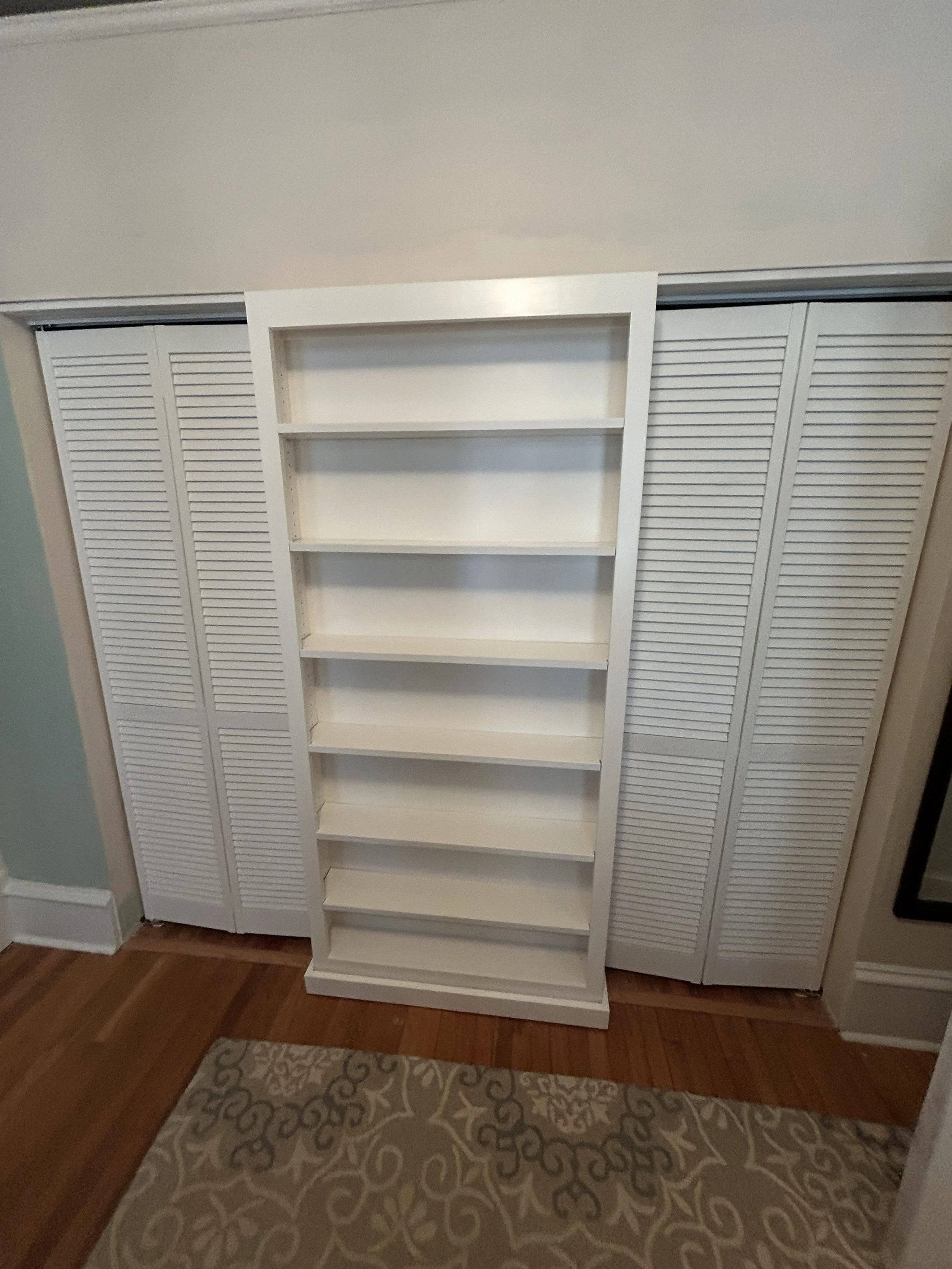 Empty white bookshelf in front of louvered closet doors in a room with wood flooring and a patterned rug.