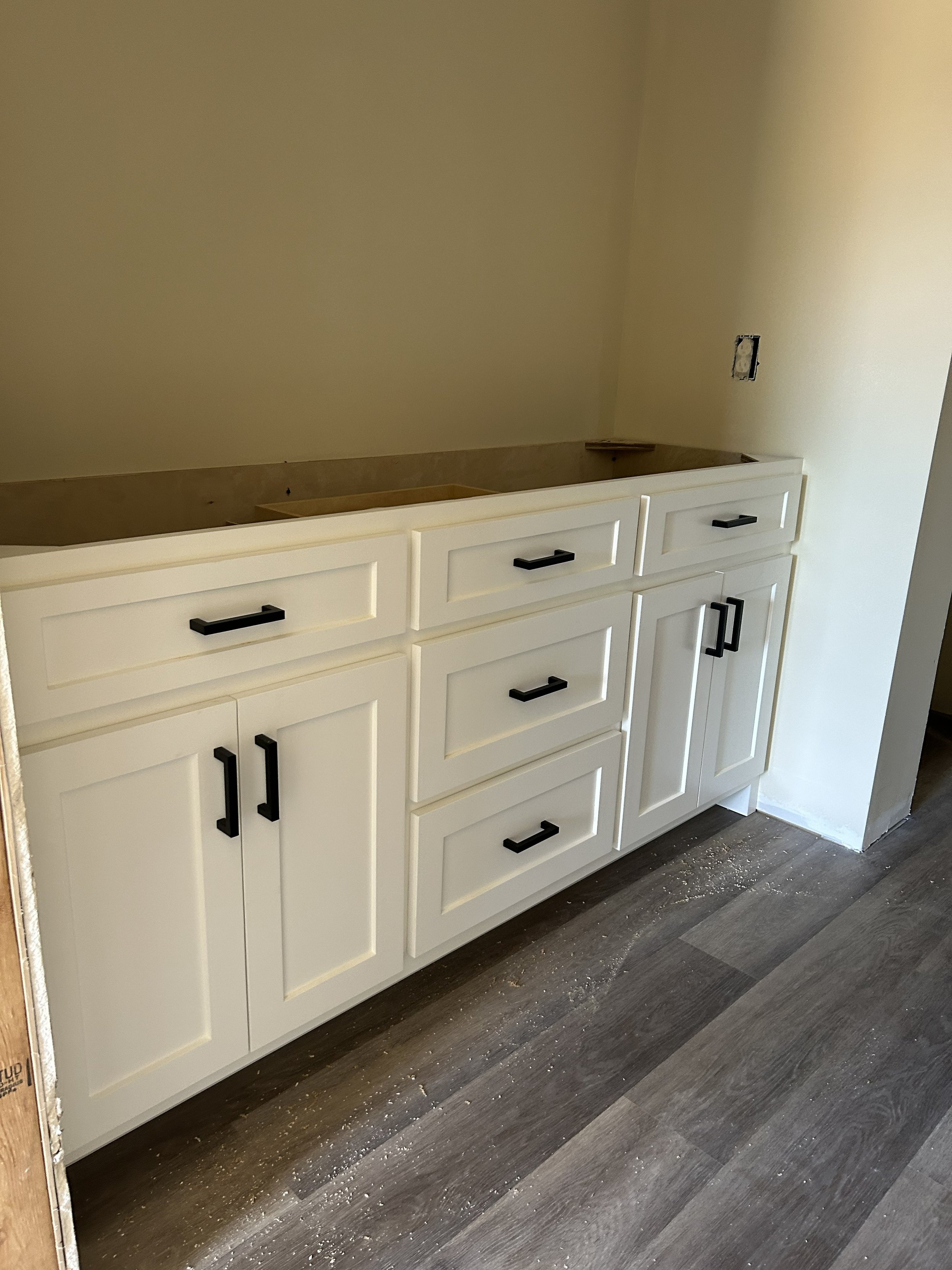 White kitchen cabinet installation in progress with black handles, with some construction dust on the floor.