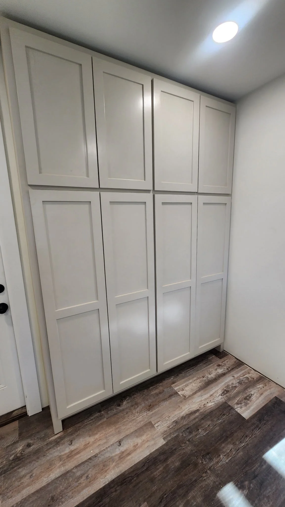 A white wooden cabinet with multiple panels in a corner of a room, set against white walls with wood-look flooring and a ceiling light.